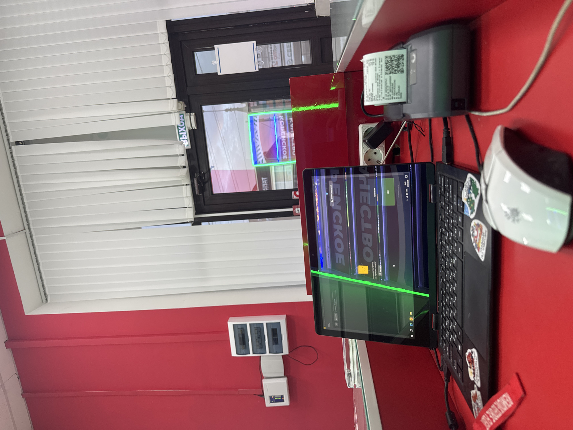 In a brightly lit room with red walls, a laptop sits open on a red desk, its screen displaying text in a foreign language and a bright green line. To the right of the laptop is a small receipt printer with a printed receipt, and a white computer mouse is partially visible in the foreground. Behind the desk, a glass partition separates the workspace from a doorway with vertical blinds. A sign above the doorway reads "ВЫХОД" (Exit) in Russian. The room appears to be a commercial space, possibly a shop or office. The presence of electrical outlets and a fuse box on the red wall suggests it is a functional workspace. The reflection in the glass indicates the scene is likely located in Yegorlykskaya, Russia, given the Cyrillic signage.