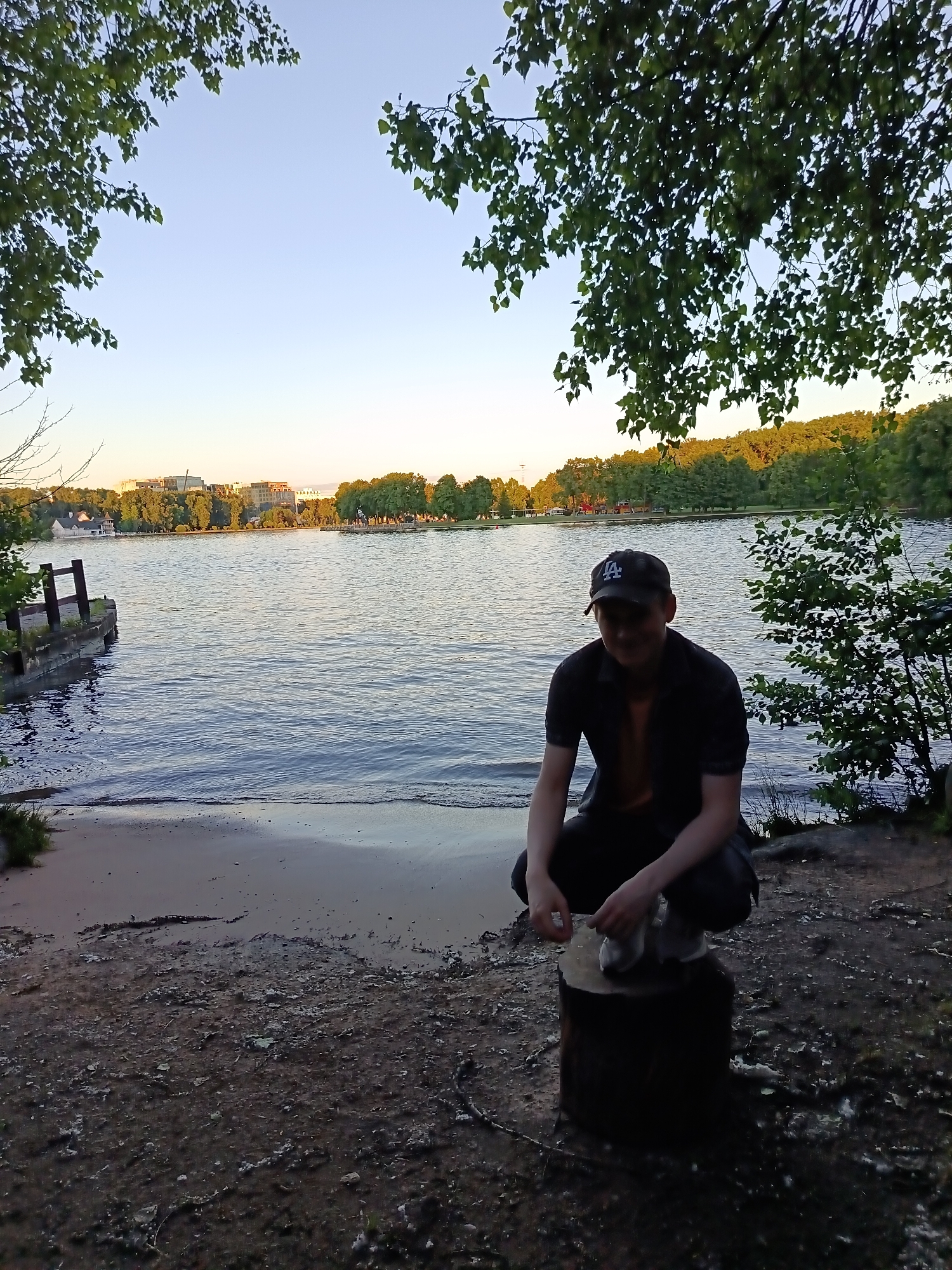 A young man wearing a black t-shirt, black pants, and a black baseball cap is crouching on a tree stump by the shore of a lake. The lake is calm and reflects the clear sky. In the background, there are trees and buildings, suggesting an urban or suburban setting. The sun is setting, casting a warm glow on the scene. The overall atmosphere is peaceful and serene. This scene is likely located at the Tsnyanskoye Reservoir, a popular recreational area in Minsk, Belarus, known for its beaches, parks, and walking trails.