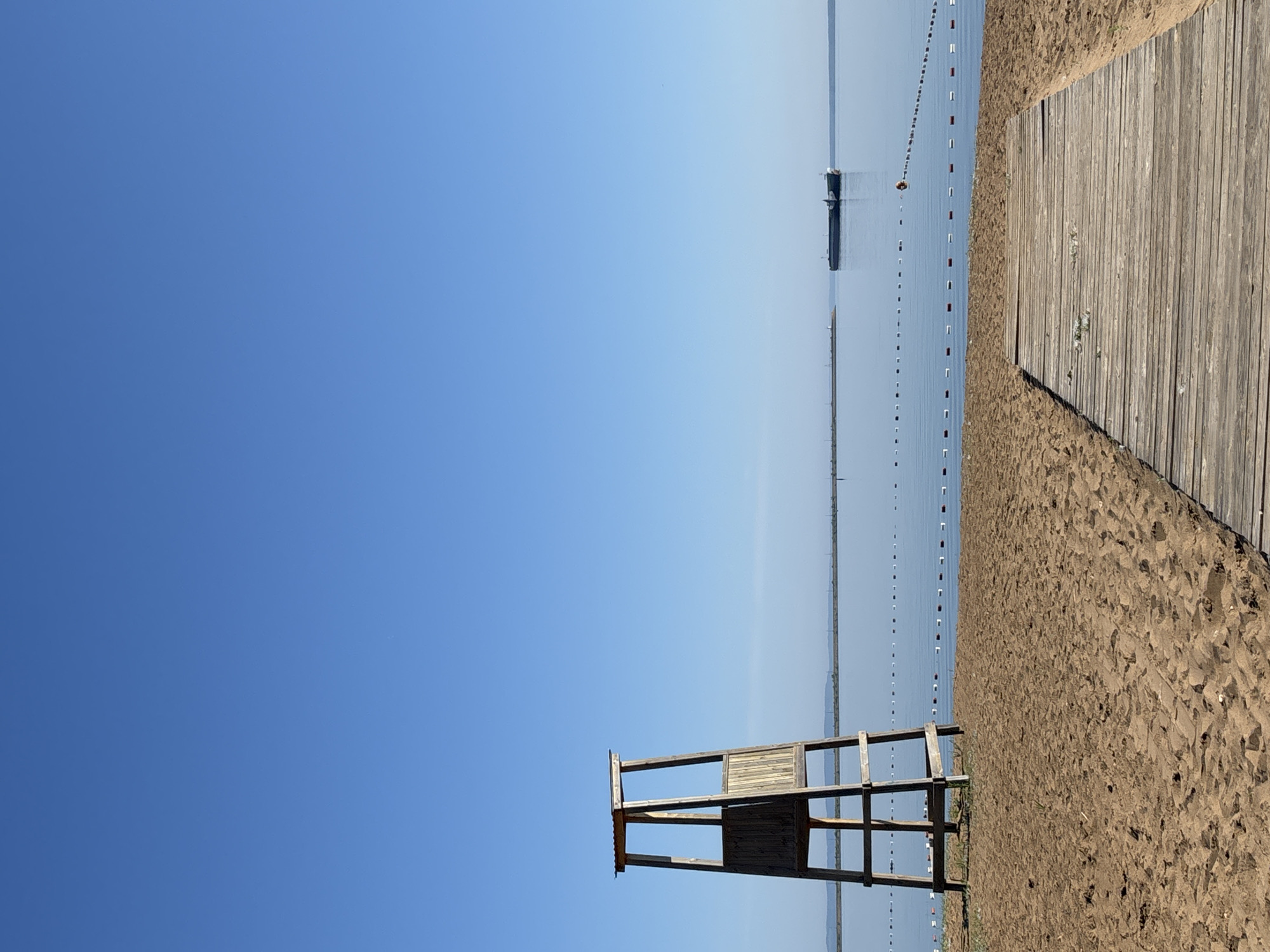 The image depicts a tranquil beach scene with a clear blue sky. In the foreground, a wooden lifeguard tower stands on the sand. A wooden walkway extends from the right side of the image towards the water. The calm water is marked by a buoy line, with a cargo ship visible in the distance on the horizon. The scene appears to be located on the Kama River embankment in Chaikovsky, Russia, a city known for its hydroelectric power station and its connection to the Kama River. The environment is serene, with no people present in the frame.