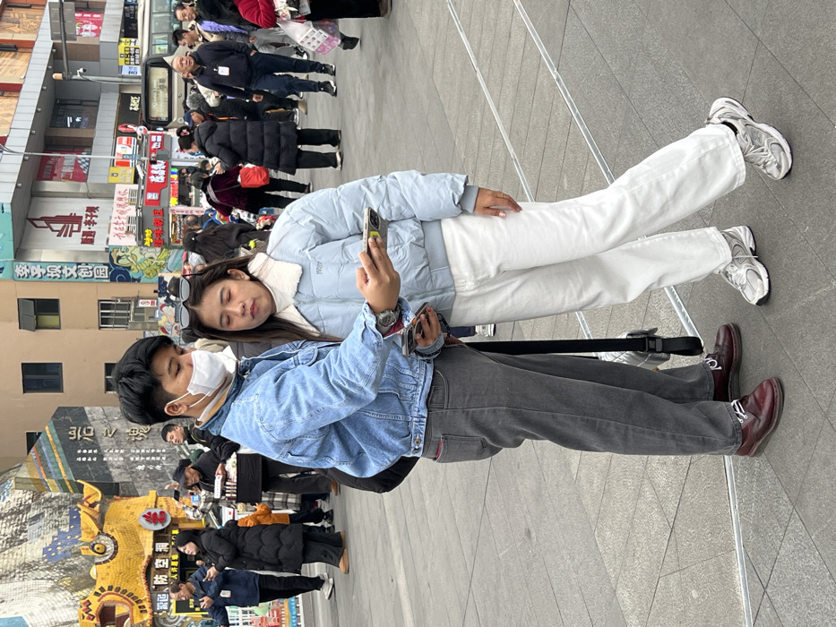 A couple stands on a gray tiled surface. The woman wears a light blue jacket, white pants, and white sneakers, resting her head on the man's shoulder. He wears a denim jacket, gray pants, brown shoes, and a white face mask while holding a phone in his hand. People and buildings fill the background.