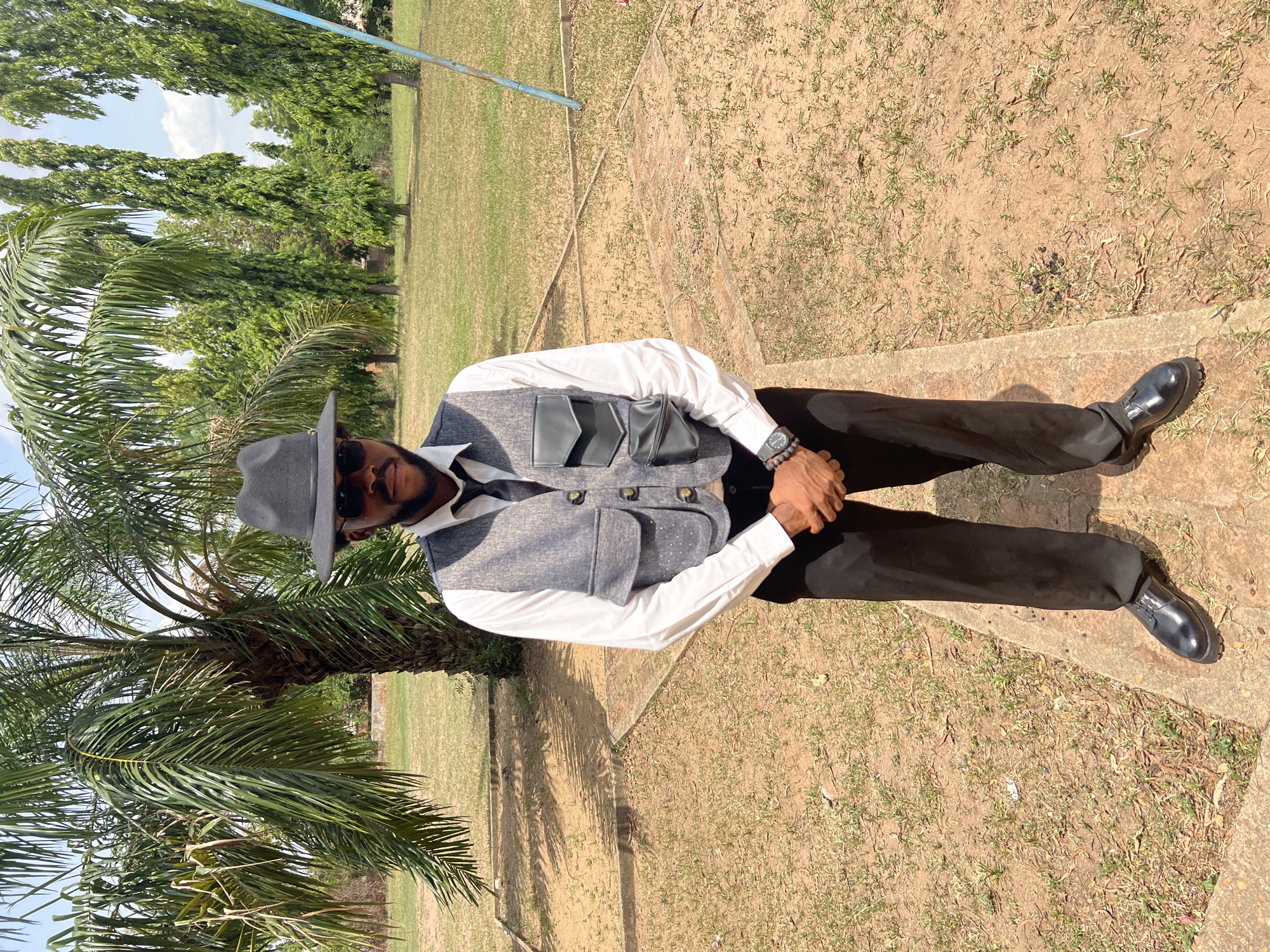 The image shows a man sitting outdoors in what appears to be a park-like setting in Markurdi, Nigeria. He is wearing a gray hat, sunglasses, a white long-sleeved shirt, a gray vest with decorative flaps, black pants, and black shoes. He has a watch on his wrist. The ground is a mix of dirt, patchy grass, and concrete. A palm tree is visible to the left of the man, and other trees are in the background under a blue sky with some clouds. A blue horizontal pole is visible in the background. The man is sitting on what looks like a set of steps or a low concrete structure.