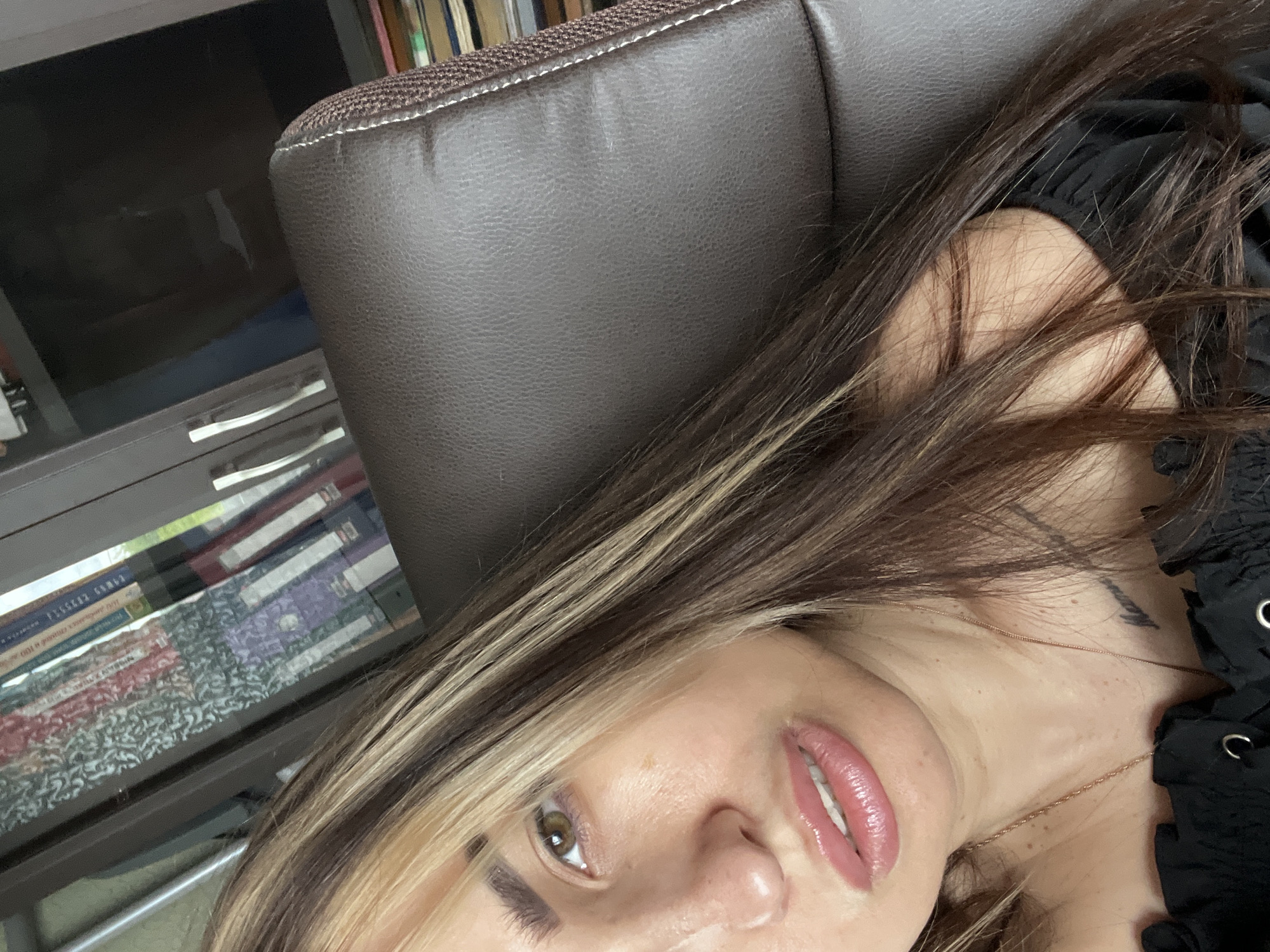 A close-up, tilted view of a person with long brown and blonde hair, light skin, and light pink lips. The person is wearing a black top. The background includes a brown leather-like chair and a shelf with books. Location: Unknown, Unknown.