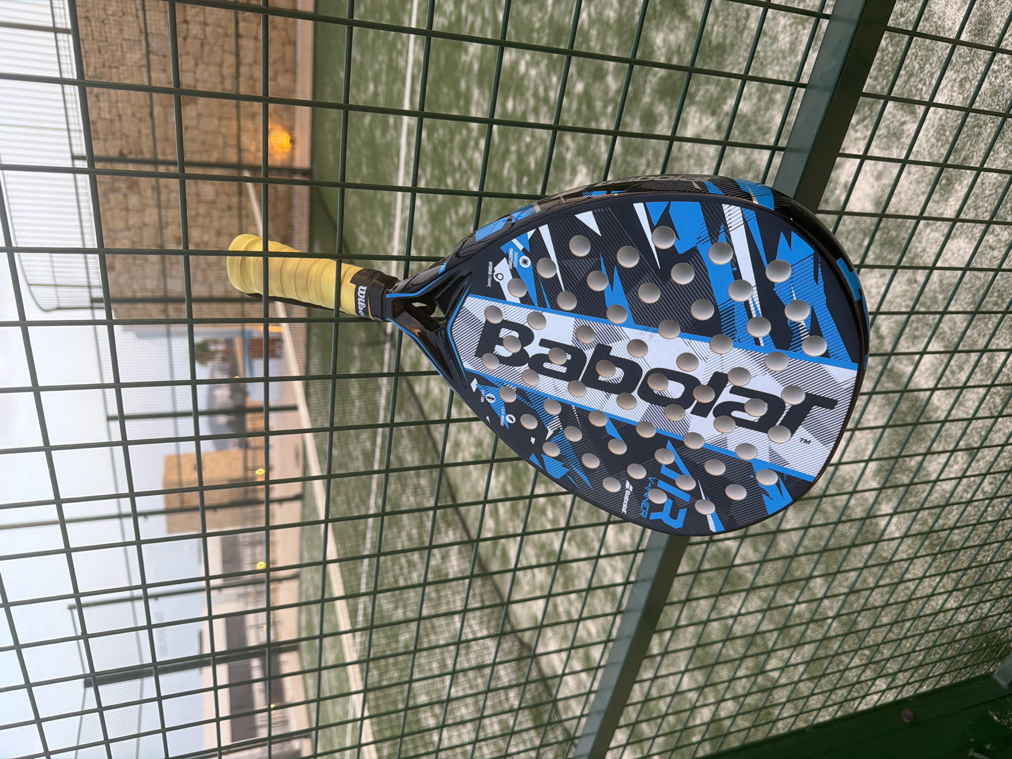 A padel racket, predominantly black with blue and white geometric designs, hangs on a green metal fence. The racket has a yellow grip and the brands "Babolat" and "Wilson" are visible. The background shows a green artificial turf padel court with white markings, and beyond that, a building with a light brown, textured facade. The scene appears to be outdoors in the city of Finestrat, Spain.