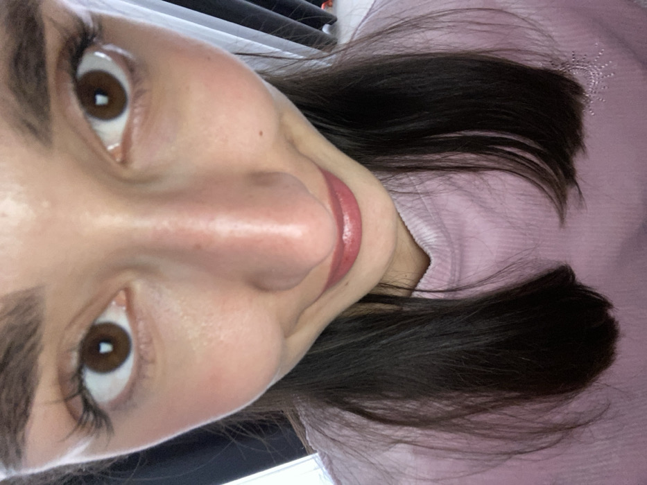 The image depicts a close-up, wide-angle portrait of an individual, presumed female, positioned indoors. The subject's face dominates the frame, looking directly into the camera with wide-open brown eyes, featuring prominent dark eyebrows and long eyelashes. A slight, closed-lip smile is indicated by the upturned corners of the mouth, which are accented with red/pink lipstick. The individual has dark brown hair that falls past the shoulders, visible on either side of the light pink, possibly ribbed, long-sleeved top. A faint white, starburst-like design is visible on the lower right chest area of the garment. The background is out of focus, showing indistinct indoor elements: a bright area, possibly a window, with a dark vertical element on the left, and a dark, vertically draped fabric, likely a curtain, on the right against a lighter surface. There are no visual cues within the image to determine a specific location within the city of Budva, Montenegro.