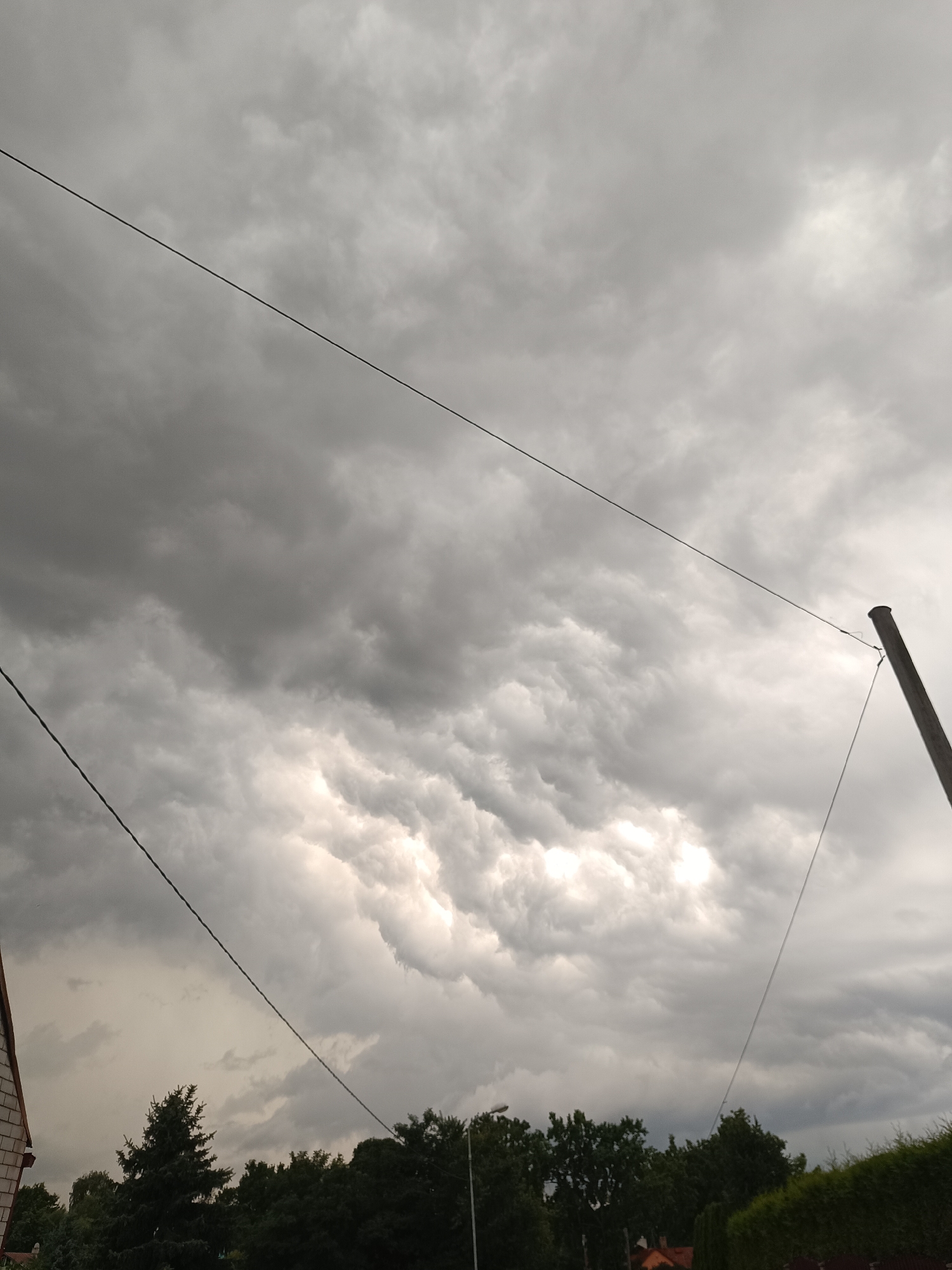 The image shows a sky filled with dramatic, dark storm clouds, with some areas of brighter light breaking through, suggesting an approaching storm or recent rainfall. The clouds have a textured, almost rippling appearance. In the foreground, silhouetted against the sky, is a line of dark green trees and foliage. A single, tall, dark pole, likely a utility pole, stands on the right side of the image. Thin wires are stretched across the sky, connecting to the pole and extending out of frame to the left. On the far left of the image, the corner of a building with a reddish-brown tiled roof and a white-brick wall is visible. The scene is set in an outdoor, possibly suburban or rural, environment, typical of areas on the outskirts of Riga, Latvia. There are no people or animals visible in the image, and no discernible actions are taking place. The overall impression is one of impending weather change.