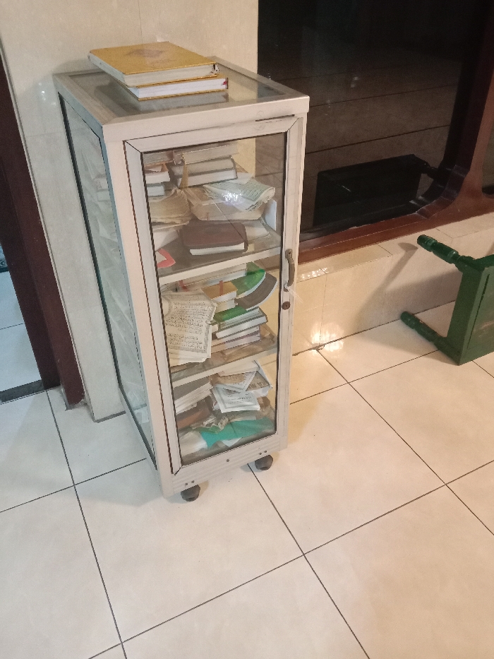 The image shows a glass-enclosed cabinet with four shelves, filled with books and documents. The cabinet has a metal frame and is sitting on four wheels. The cabinet is located in a building with a tiled floor. The cabinet is near a door with wood trim and another door with dark glass. The floor is tiled in a cream-colored square pattern. The cabinet is also near a brown wooden door with a black frame and an object that appears to be a bench with a dark green seat. There are no people visible in the image. The location of the image is a building in the city of Tugu, Indonesia, based on the provided coordinates of -6.976 latitude and 110.331 longitude.