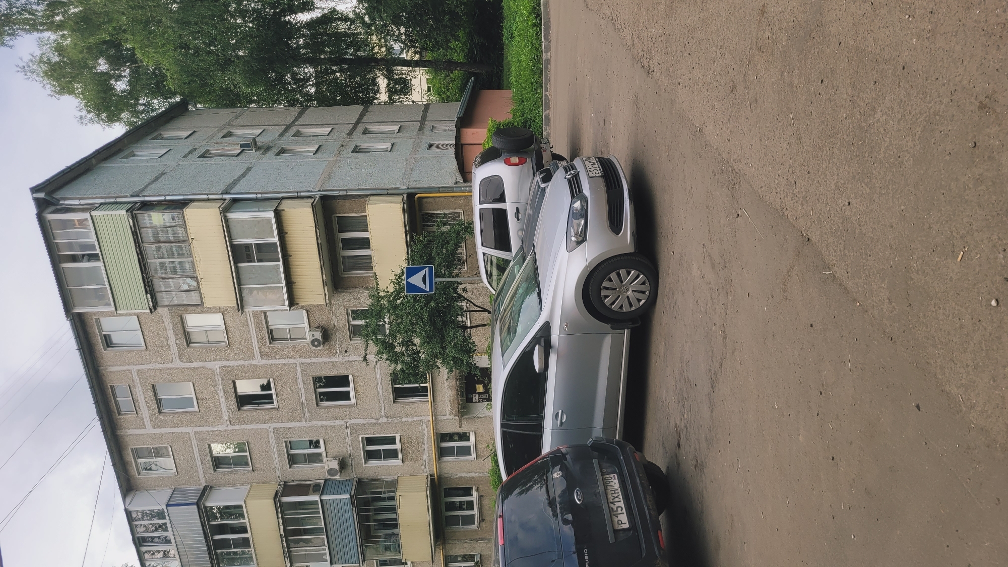 A medium shot shows a silver Volkswagen Polo sedan parked on a dirt and asphalt lot next to a grey, five-story apartment building. The building has multiple balconies with glass enclosures and is surrounded by green trees. A black Ford Fusion is parked to the left of the Polo, and a silver SUV with a spare tire mounted on the rear is parked behind the Polo. A blue and white road sign with a white triangle inside a blue square is visible between the apartment building and the cars. The sky is overcast.