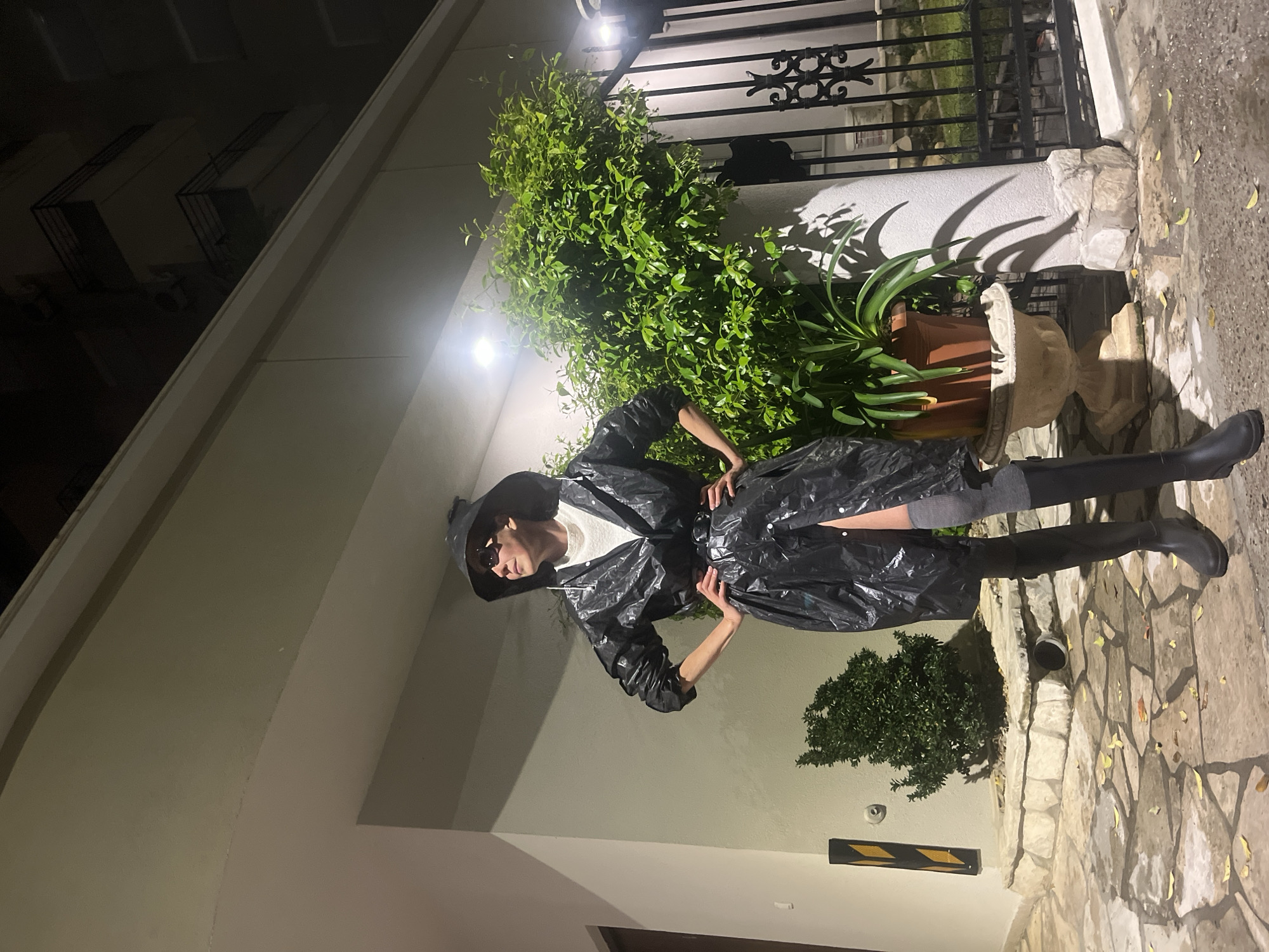 A full-body, portrait-oriented photograph, taken at night, features an individual posing outdoors. The person stands with hands on hips, facing slightly left, wearing a distinctive black, glossy raincoat or trench coat, a white t-shirt, black rain boots, and visible grey socks. A black hat and dark sunglasses complete the ensemble, obscuring the person's facial features.
The setting is an outdoor paved area, possibly a pathway or entrance next to a building. The ground is composed of irregularly shaped stone pavers, with a few light-colored fallen leaves scattered. On the right, a large potted plant, possibly an agave, rests on an ornate stone pedestal, flanked by a black wrought-iron fence or gate. Lush green foliage, including a prominent leafy bush, fills parts of the background. The scene is illuminated by strong artificial spotlights, casting prominent shadows from the plants and the figure onto the light-colored building wall that forms much of the background. A small black and yellow striped hazard marker is visible near the bottom left. The deliberate pose and unique attire suggest a fashion-oriented or stylized shot. No visible text is present in the image.