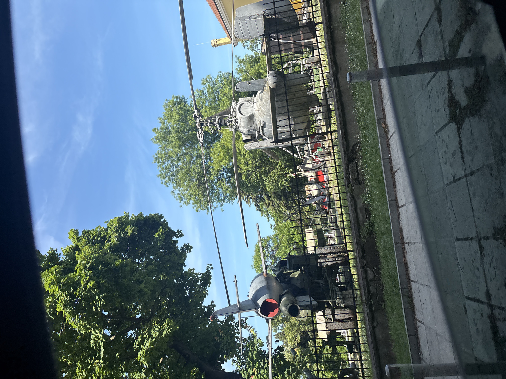 The image displays an outdoor museum exhibit featuring a grey Kamov Ka-25 helicopter on the right and a grey P-15 Termit anti-ship missile mounted on a wheeled launcher on the left. Both military artifacts are situated behind a black metal fence, which stretches across the mid-ground of the scene. The background consists of dense green trees under a clear blue sky with minimal white clouds. A yellow building with a red-tiled roof is partially visible behind the helicopter on the far right. The immediate foreground shows a paved concrete sidewalk, with a concrete curb separating it from a strip of grass and small white wildflowers adjacent to the fence. A grey cylindrical pole stands upright on the sidewalk in the lower right. The image appears to be taken from inside a vehicle, evidenced by a dark bar obscuring the top portion and a reflective surface across the bottom. No people are visible. This scene is identified as part of the outdoor exhibition at the Naval Museum (Военноморски музей) in Varna, Bulgaria, located within the Sea Garden.