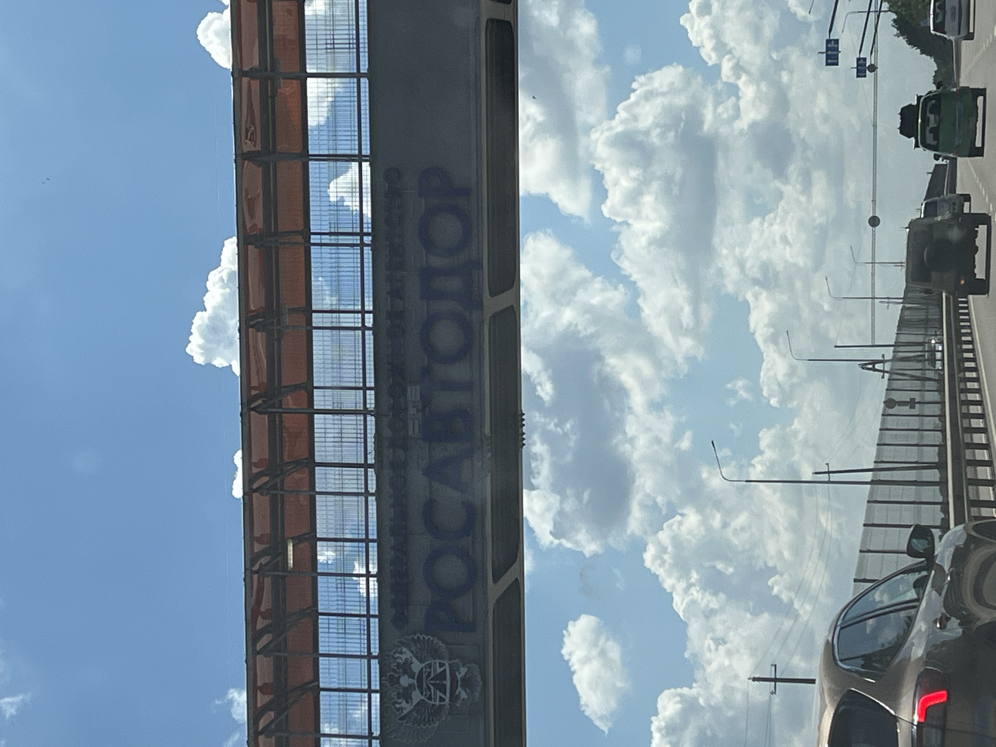 This image, taken in Belozyorny, Russia, depicts a scene from a moving vehicle, likely a car, on a highway on a cloudy day. The perspective is from inside the vehicle, looking upwards and outwards.
The main subject is a large, elevated structure, possibly an overpass or a part of a bridge, running vertically through the frame. This structure is metallic, with a dark gray main beam and a latticed, reddish-orange covering on its upper section, possibly a roof or protective barrier. Large, blue letters are painted on the side of the structure, spelling out "РОСАВТОДОР" (ROSAVTODOR). Below the text, there is an emblem featuring a double-headed eagle with a shield containing a St. Andrew's cross.
The sky is filled with dramatic, fluffy white cumulus clouds against a bright blue sky, suggesting it is daytime, likely mid-afternoon given the intensity of the light. The weather appears warm and potentially humid.
Other objects visible include traffic. Several vehicles are on the highway ahead, including a green truck and a military-style vehicle. The perspective from the camera's car shows the rear of a dark-colored sedan in the foreground, its taillight illuminated. Streetlights and traffic signals are also visible along the roadside.
The scene conveys a sense of travel and movement through a somewhat urban or suburban environment, characterized by infrastructure and vehicular activity under a dynamic sky. There are no people clearly visible as subjects, but the presence of vehicles implies human activity. The overall impression is one of a typical summer day's travel.