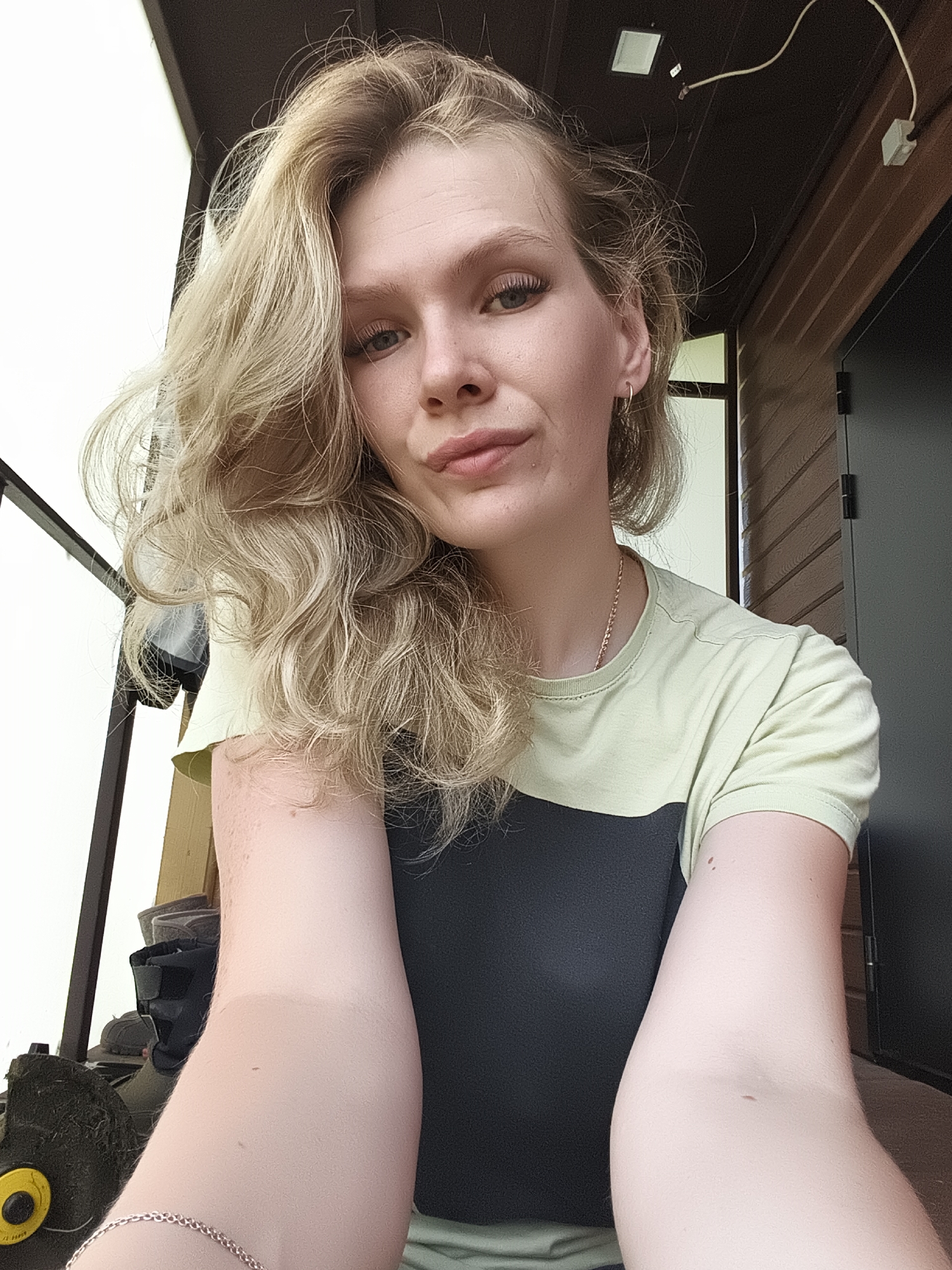A young woman with blonde, wavy hair and light-colored eyes is taking a selfie. She is wearing a t-shirt with a light green top half and a dark gray bottom half. She has a small gold necklace on and a thin gold bracelet on her left wrist. Her left arm is extended towards the camera, and her right arm is bent, with her hand resting on her chest. Her expression is neutral, with her lips slightly pursed.
The background appears to be outdoors, possibly on a porch or patio. There are wooden planks on the ceiling and a wall made of wooden siding. A light fixture is visible on the ceiling, and a wire runs from it to a junction box. To the left of the woman, there is a glimpse of some outdoor equipment, including what looks like a lawnmower. The environment suggests a residential or recreational area. The image was taken in Русский Вожой, Russia.