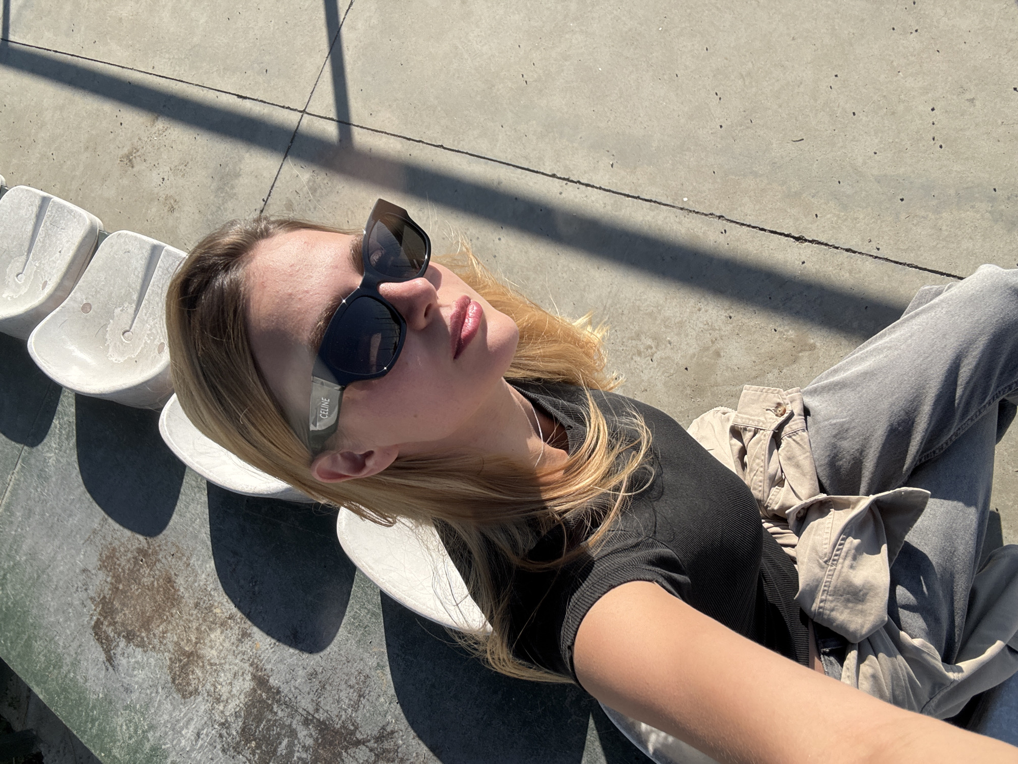 A young adult female poses for a selfie while sitting in the bleachers of Dinamo Arena in Tbilisi, Georgia. She is wearing dark sunglasses, a black top, and light-colored jeans with a beige jacket draped on her lap. The seating area is concrete and white plastic chairs. Sunlight casts shadows across the scene.