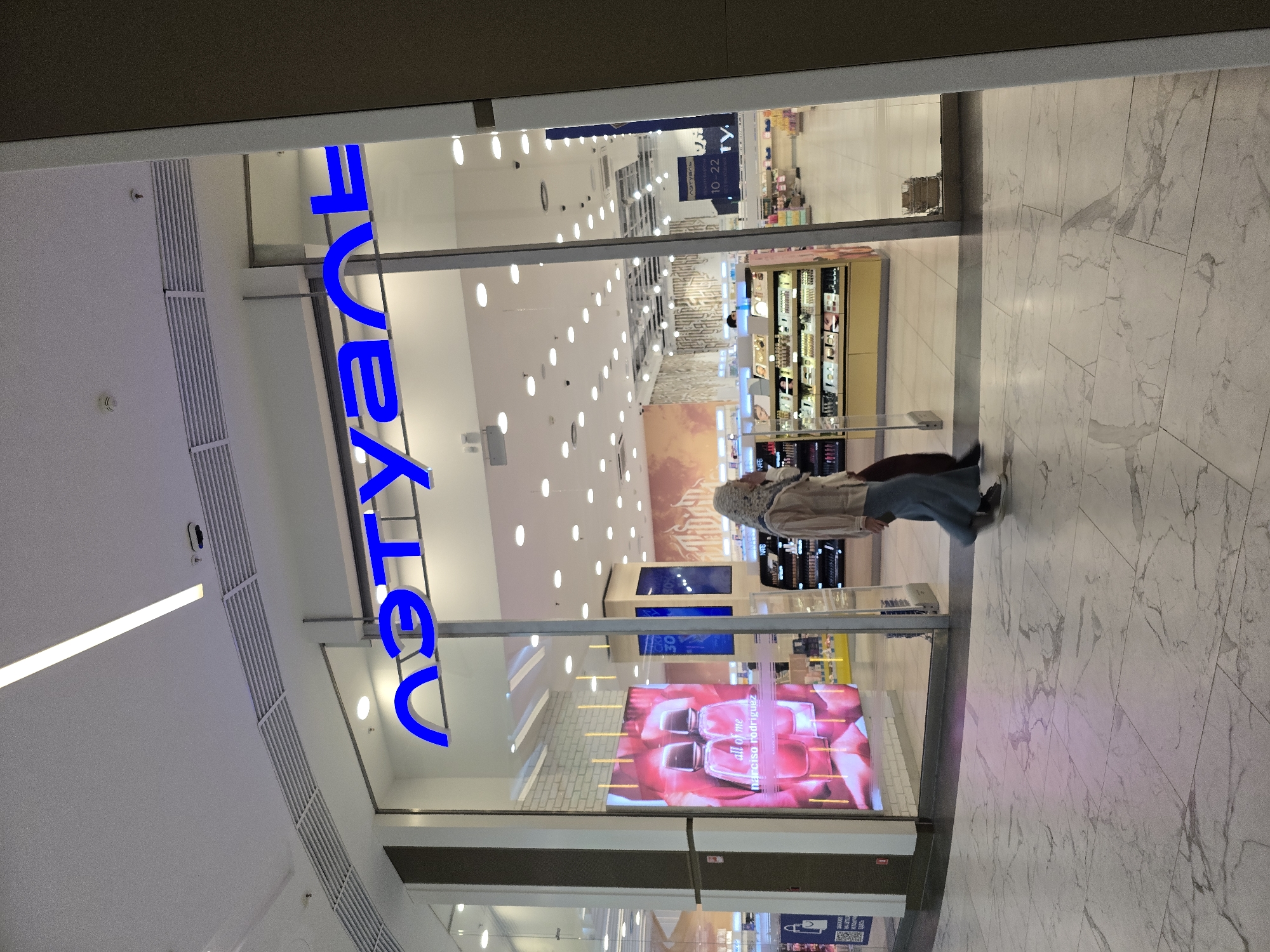 A street-level, full shot depicts a woman walking past a store called "Yves Rocher." The store is located in Grozny, Russia, evidenced by the Cyrillic letters spelling "Yves Rocher." The woman is wearing a hijab, a tan jacket, and blue jeans. She is walking on a white marble floor. The storefront has large glass windows that display cosmetics and perfume advertisements. The interior of the store is brightly lit with many overhead lights. The exterior of the store is in a mall.