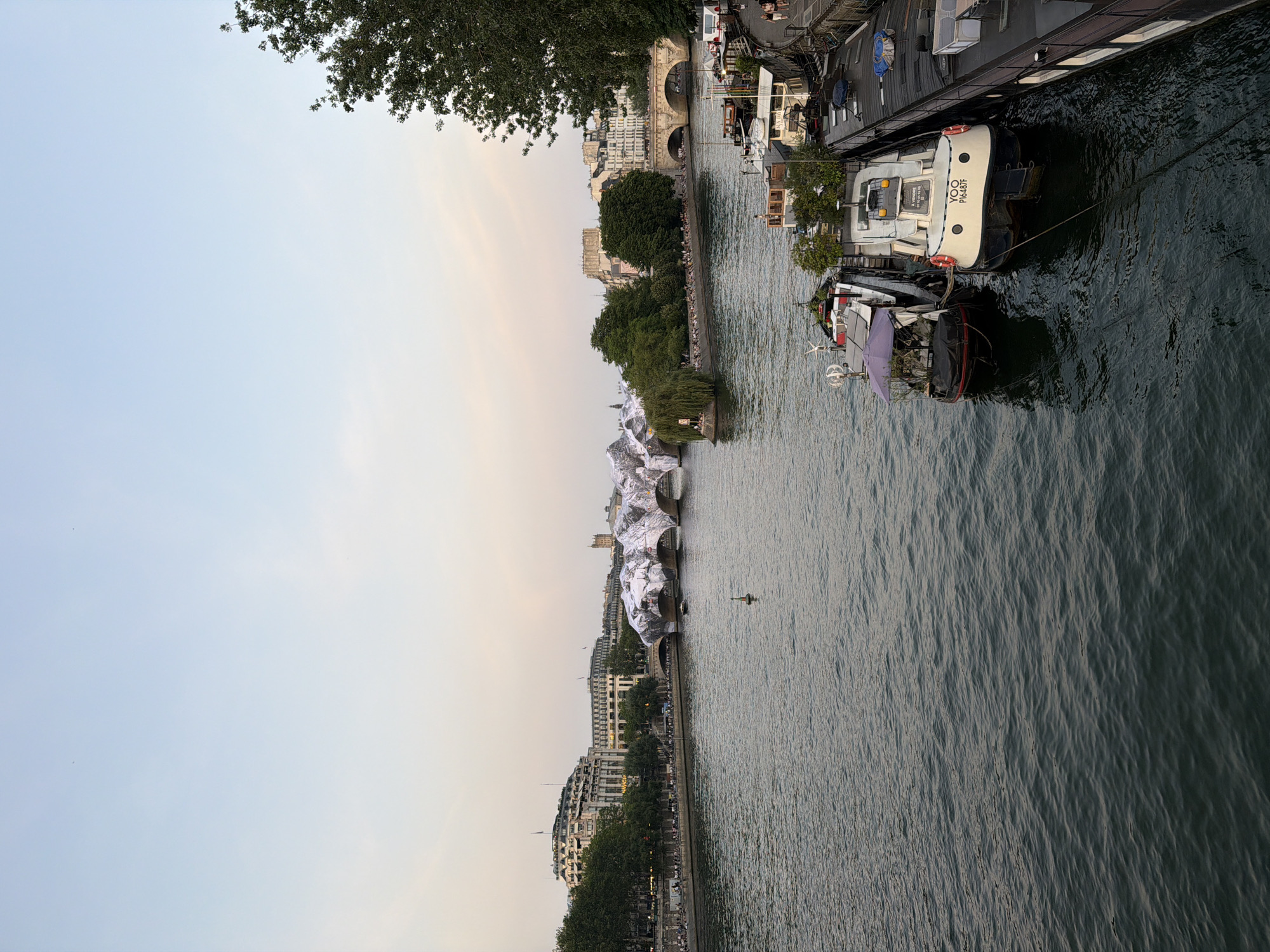 This image captures a view of the Seine River in Paris, France, with several boats docked along the riverbanks. The river's surface is slightly rippled, suggesting a gentle breeze. On one side of the river, a line of boats is covered in what appears to be a temporary art installation, likely involving white fabric or material draped over the boats. People are visible along the riverbanks, suggesting a leisurely atmosphere. Buildings line the riverbanks, adding to the Parisian cityscape. In the background, a bridge can be seen, providing a sense of depth and location within the city. A tree is in the upper corner of the shot. The light in the image suggests either sunrise or sunset, casting a soft glow over the scene.