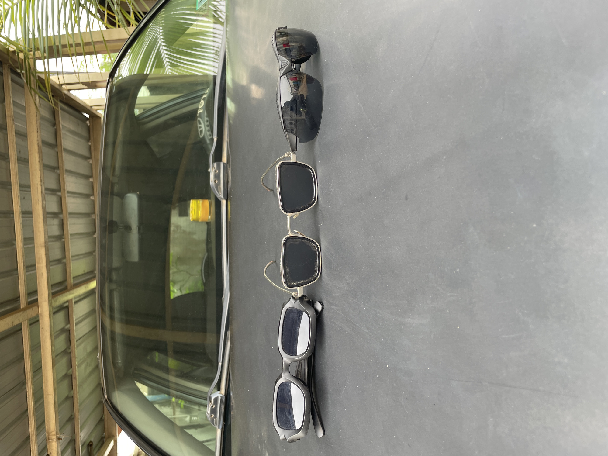 The image shows three pairs of sunglasses resting on the hood of a vehicle. The sunglasses are arranged vertically in the center of the frame. The vehicle is parked in an area with corrugated metal walls and a palm tree visible overhead. The location is Akwanga, Nigeria.