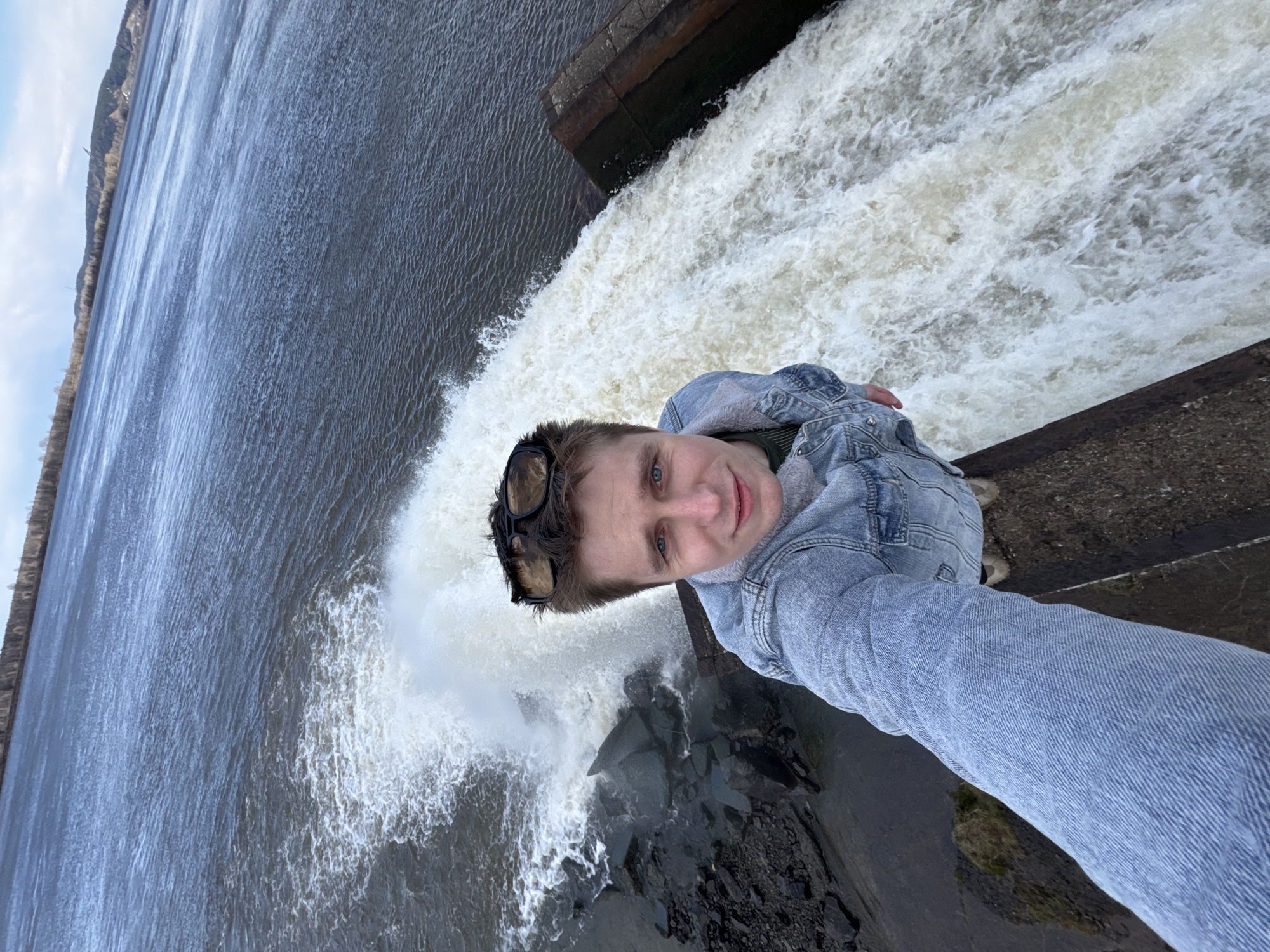A young man takes a selfie-style photograph beside a powerful waterfall or dam spillway, with water vigorously cascading downwards. He has light-colored hair, blue eyes, and wears a light blue denim jacket over a darker shirt, with black-framed sunglasses pushed onto his head. He looks directly at the camera with a slight smile.
The man stands on a dark, concrete-like structure, part of the dam or spillway. To his left, a vast expanse of water, appearing as a river or reservoir, stretches towards the horizon, showing rippled surfaces. In the background, a tree-covered hillside runs along the far bank under a partly cloudy sky. Below the man, the water plunges, creating a wide, frothy, white waterfall effect that continues into the dark, rocky area below the dam. The scene captures a daytime moment, likely in Noviy, Russia, showcasing an impressive natural and engineered landscape. No visible text is present in the image.