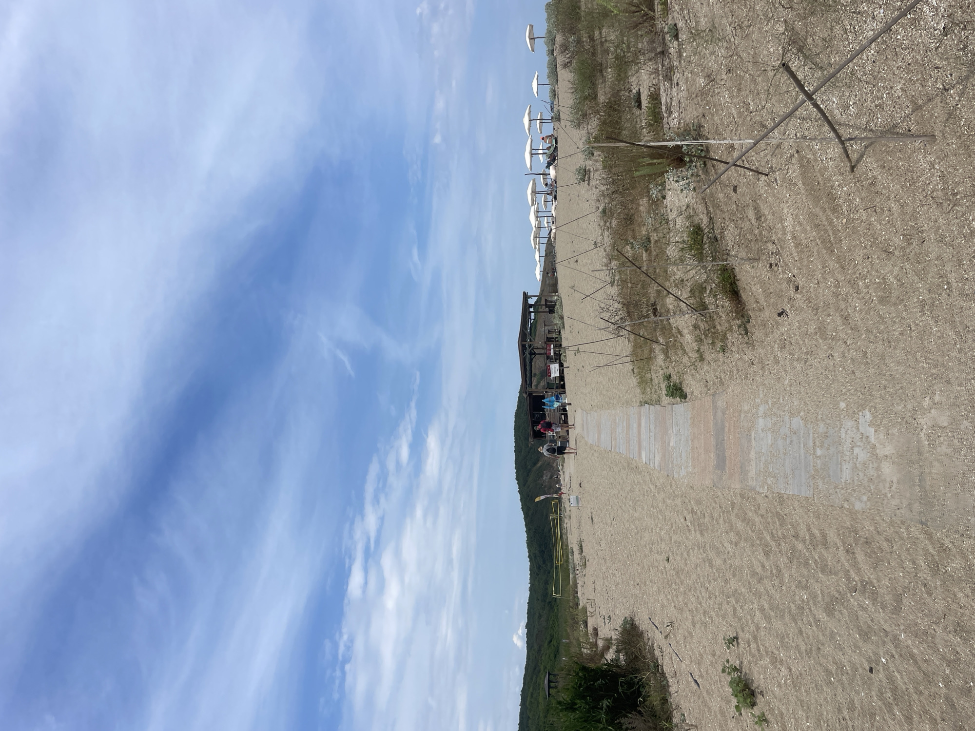 The image shows a sandy beach with a wooden boardwalk leading towards the water. In the distance, there are several white umbrellas set up on the sand, suggesting a beach resort or public beach area. A shaded structure, possibly a beach bar or cafe, is visible behind the umbrellas. Green hills form the background, covered in dense vegetation. The sky is a light blue with scattered white clouds. A few people are walking on the boardwalk, and some are seated under the umbrellas. There is a volleyball net set up on the beach to the left of the boardwalk. The overall scene depicts a sunny day at a beach in Sinemorets, Bulgaria.