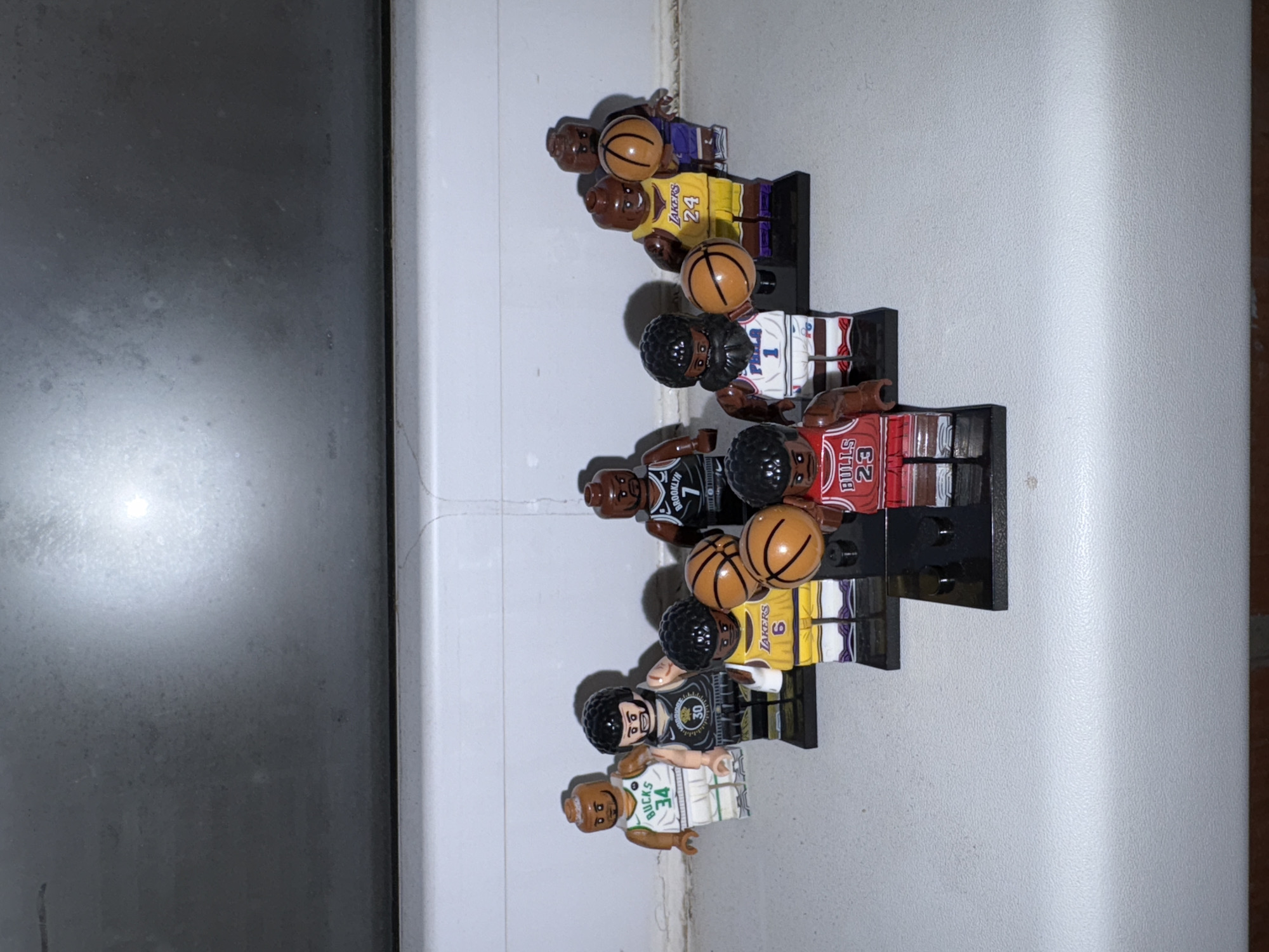 The image shows a collection of seven Lego minifigures dressed in basketball uniforms and holding basketballs, arranged in a slightly tilted vertical stack on a light-colored surface next to a dark gray surface. The minifigures represent different NBA players, with visible jersey numbers including 24 (Lakers), 1 (unknown team), 23 (Bulls), 7 (Nets), 6 (Lakers), 30 (Warriors), and 34 (Bucks). The background appears to be a windowsill. No information suggests the location is in Izhevsk, Russia.