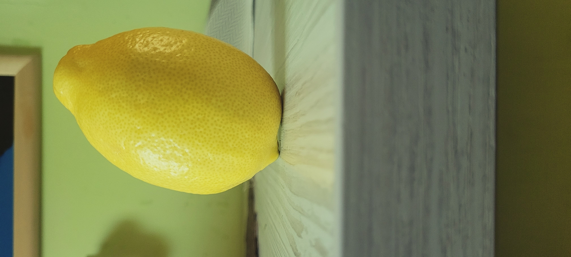 A single, bright yellow lemon rests on a light-colored wooden surface, angled slightly towards the viewer. The lemon has a textured rind and a slight point at its upper end. Behind the lemon, the background is a soft, pale green. The foreground is dominated by a blurred, grey, horizontal surface, suggesting a table or shelf in front of the wooden surface on which the lemon sits. The lighting appears to be from the side, casting a subtle shadow to the left of the lemon. The image is framed from a low angle, emphasizing the lemon as the central subject.