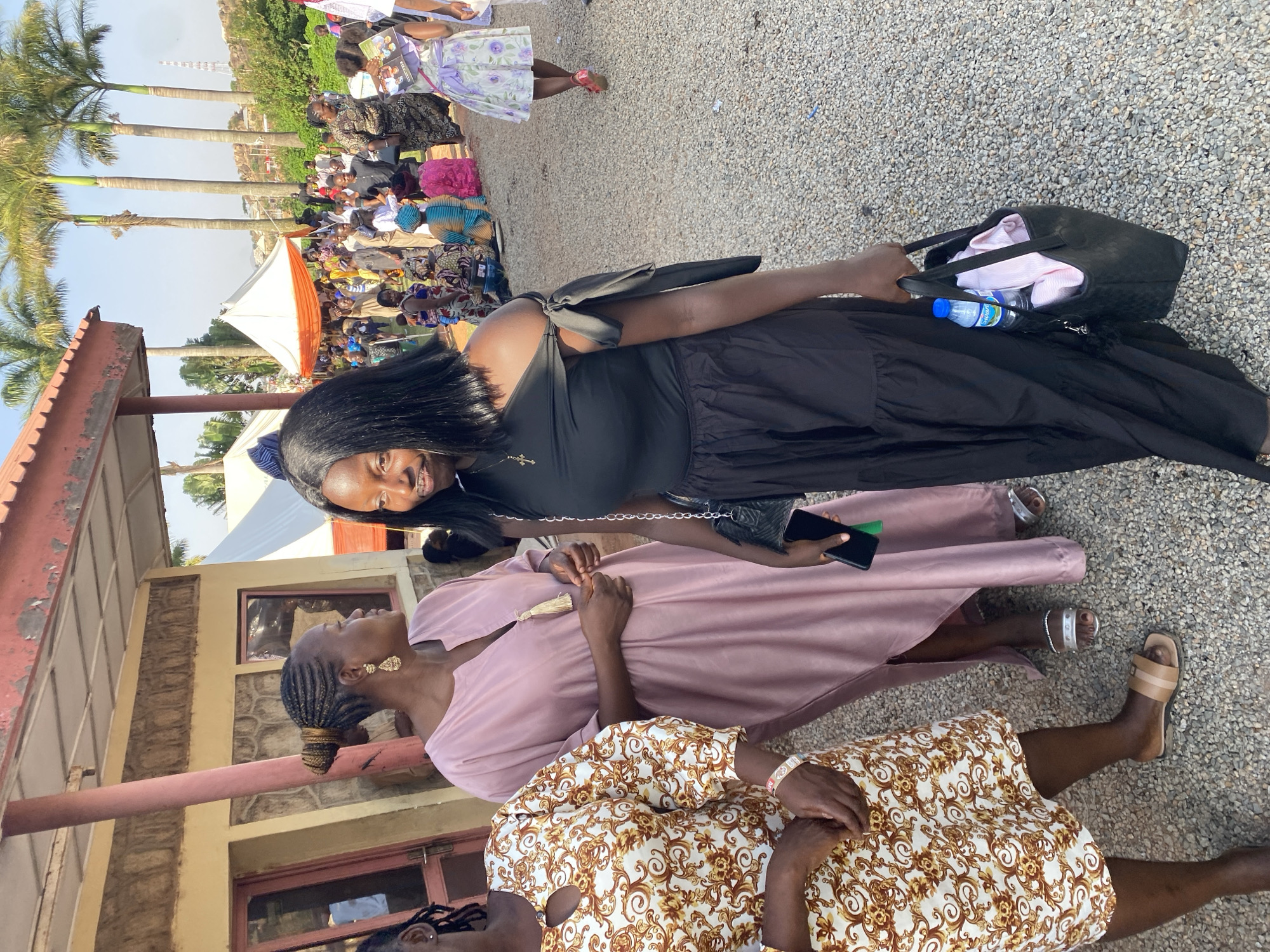 The scene appears to be at an outdoor gathering in Jos, Nigeria, possibly a social or religious event. Several people are present, and there is a crowd visible in the background. In the foreground, three women are prominent. One woman is in a black dress, holding a handbag and a phone. Another woman wears a pink dress. The third woman has on a dress with a white and brown floral pattern. Some of the attendees sit under a tent. Palm trees and buildings are in the background. The setting suggests a warm, sunny day.