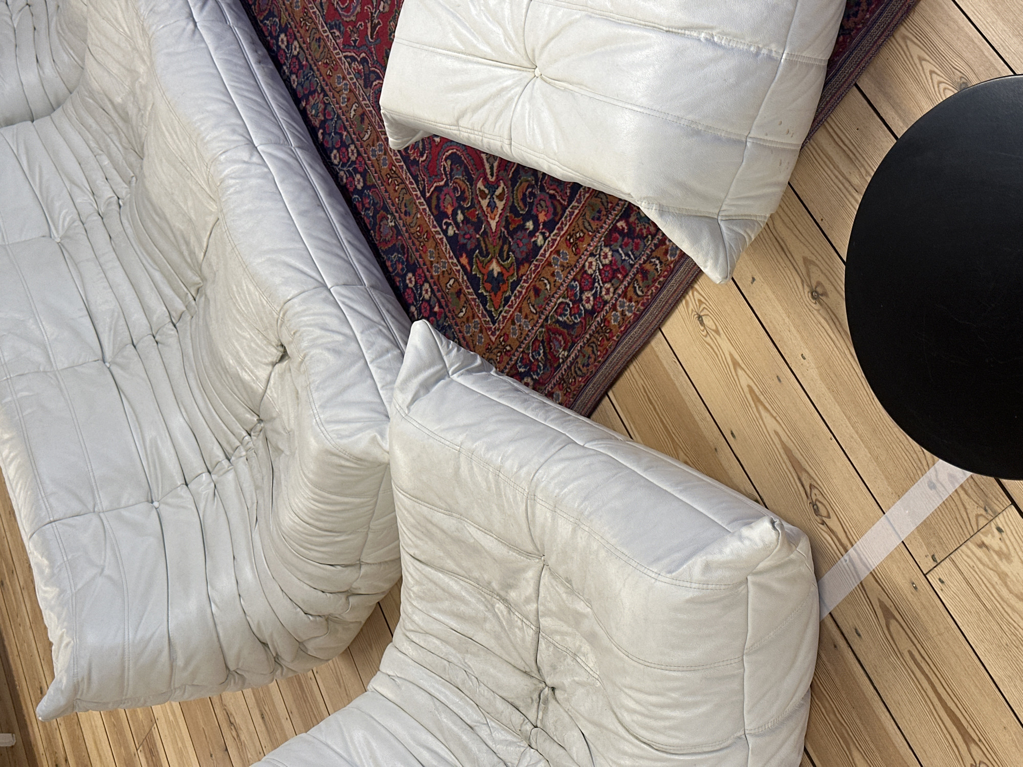 The image shows an interior scene with a partial view of a white leather Togo sofa and a matching ottoman positioned on a patterned rug. A small black, round table sits on a wooden floor adjacent to the rug and furniture. The scene is devoid of people. The setting does not clearly indicate a location in London, United Kingdom.