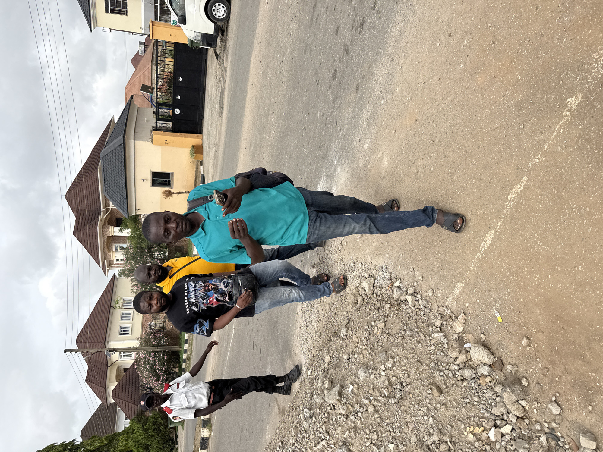 A group of four men are standing on the side of a paved road with a rough gravel shoulder in what appears to be a residential area of Abuja, Nigeria. A man in a light blue shirt is holding a cell phone. Adjacent to him is a man in a yellow shirt. Another man in a black T-shirt with a graphic print stands next to the man in the yellow shirt and carries a black shoulder bag. A fourth man, wearing a white shirt and black pants, stands slightly apart from the group. Buildings with reddish-brown roofs are visible in the background. The sky is overcast. Power lines run across the top left of the frame. A vehicle is parked on the street near one of the houses.