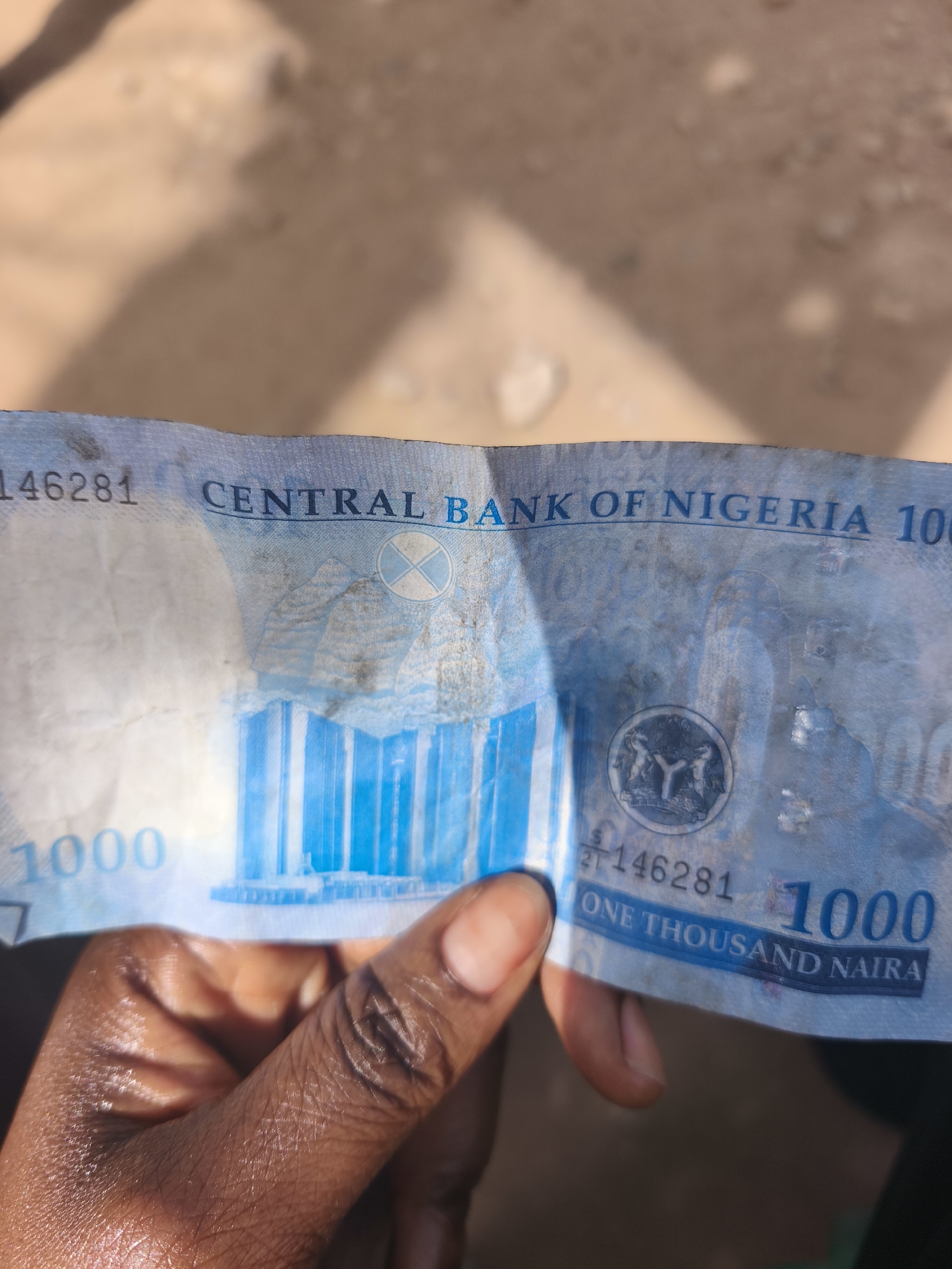 A close-up, eye-level shot depicts a person's dark-skinned hand holding a Nigerian 1000 Naira banknote outdoors under bright sunlight. The banknote is predominantly blue, slightly crumpled, and shows signs of use, including some creases and dirt marks.
The front of the note clearly displays "CENTRAL BANK OF NIGERIA" across the upper center. The denomination "1000" is visible in the bottom left and right corners, and "ONE THOUSAND NAIRA" is printed along the bottom edge. A partial serial number, "L146281," appears on both the left and right sides. In the center, a large blue graphic of a building with multiple columns, likely representing the Central Bank of Nigeria headquarters, is prominent. Above this, a circular watermark or security feature containing a cross-like symbol is faintly visible. To the right, a circular emblem, possibly Nigeria's coat of arms, shows two horses supporting a central Y-shaped symbol.
The hand gripping the note has distinct skin texture, with visible lines and folds on the fingers and palm. The background is blurred, revealing light brown, sandy, or dusty ground, consistent with an outdoor setting in a location like Jos, Nigeria. The harsh, direct lighting suggests it is midday or early afternoon, casting strong highlights and subtle shadows on the banknote and the hand.