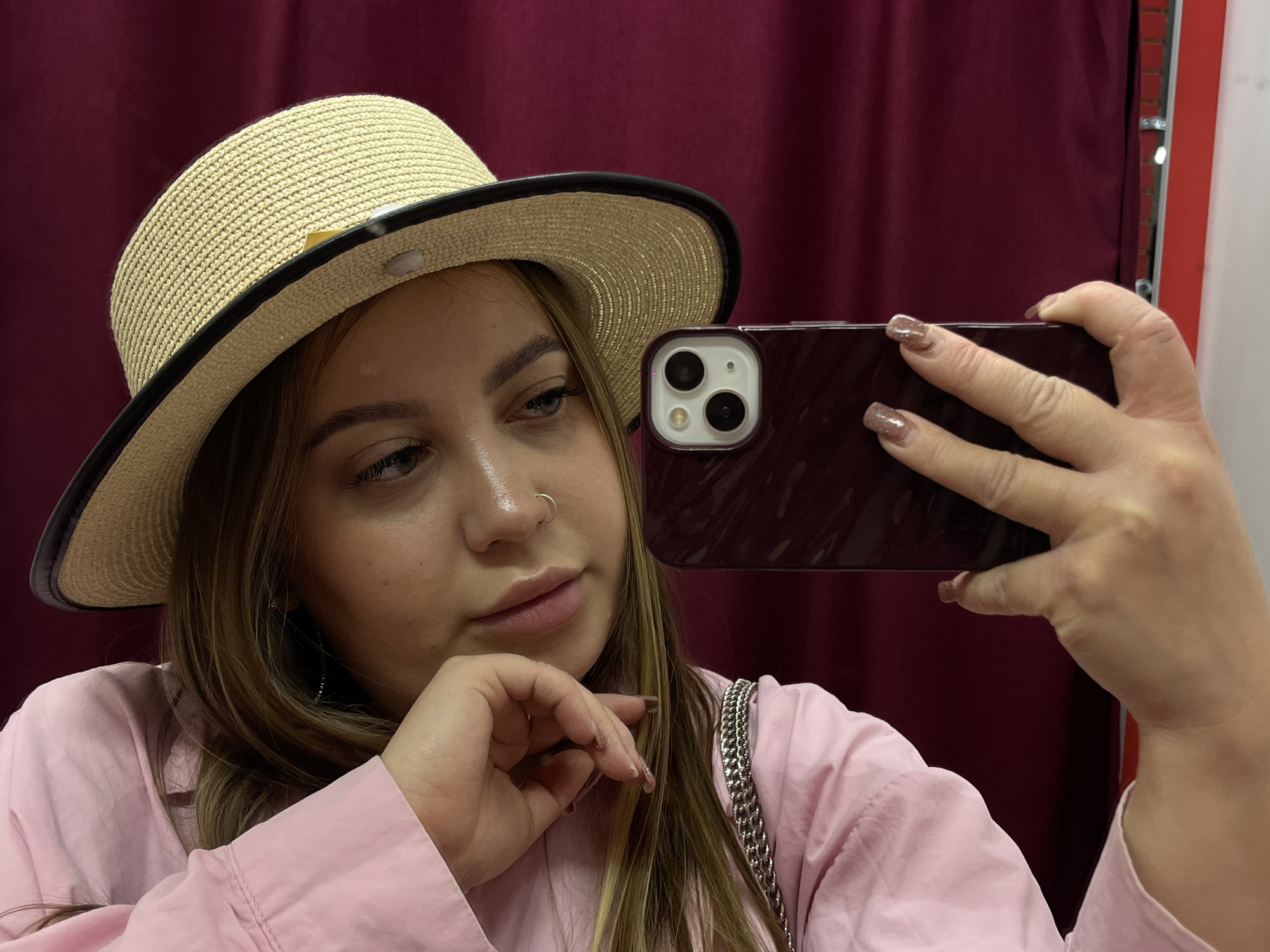 A young woman with blonde hair, wearing a straw hat with a black brim and a pink button-up shirt, is taking a selfie with a maroon-colored smartphone. She is holding the phone in her right hand, and her left hand is near her chin. She has a nose ring. The background features a burgundy curtain and a partially visible white wall. The location is unknown, and there is no indication of Novopokrovskaya, Russia.