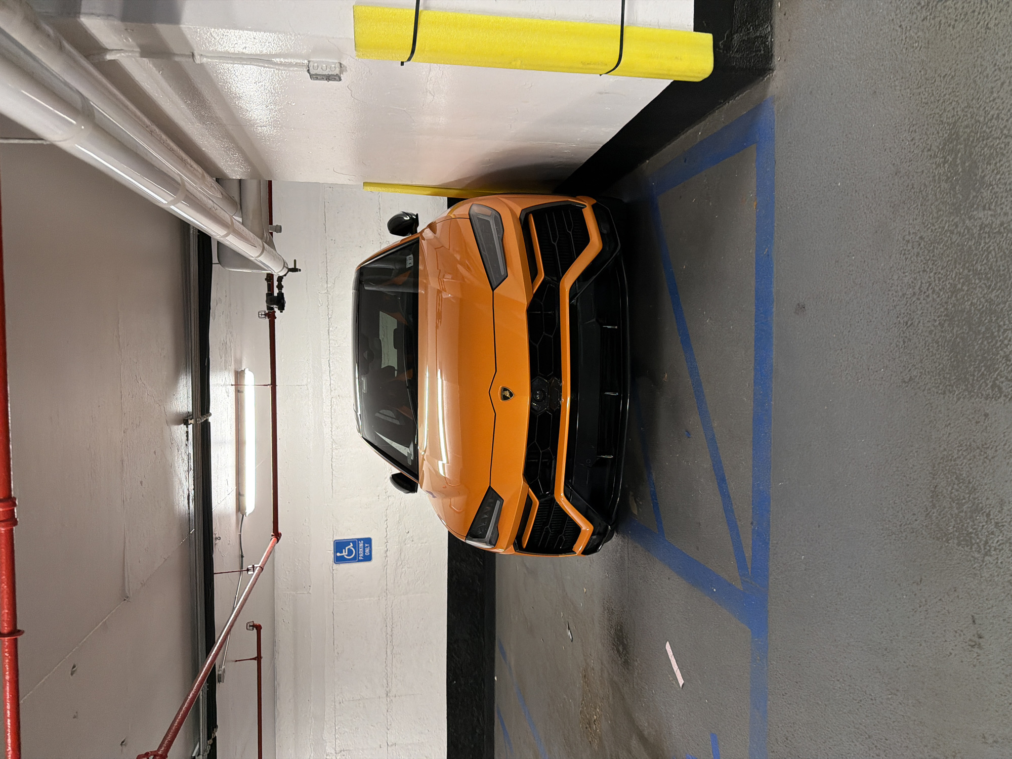 An orange Lamborghini Urus is parked in a handicapped parking spot in an indoor parking garage. The garage has white walls and ceiling with red pipes and fluorescent lighting. Blue lines mark the parking spaces, and a blue handicapped parking sign is visible on the wall. There are yellow padded bollards on the right side of the parking space.