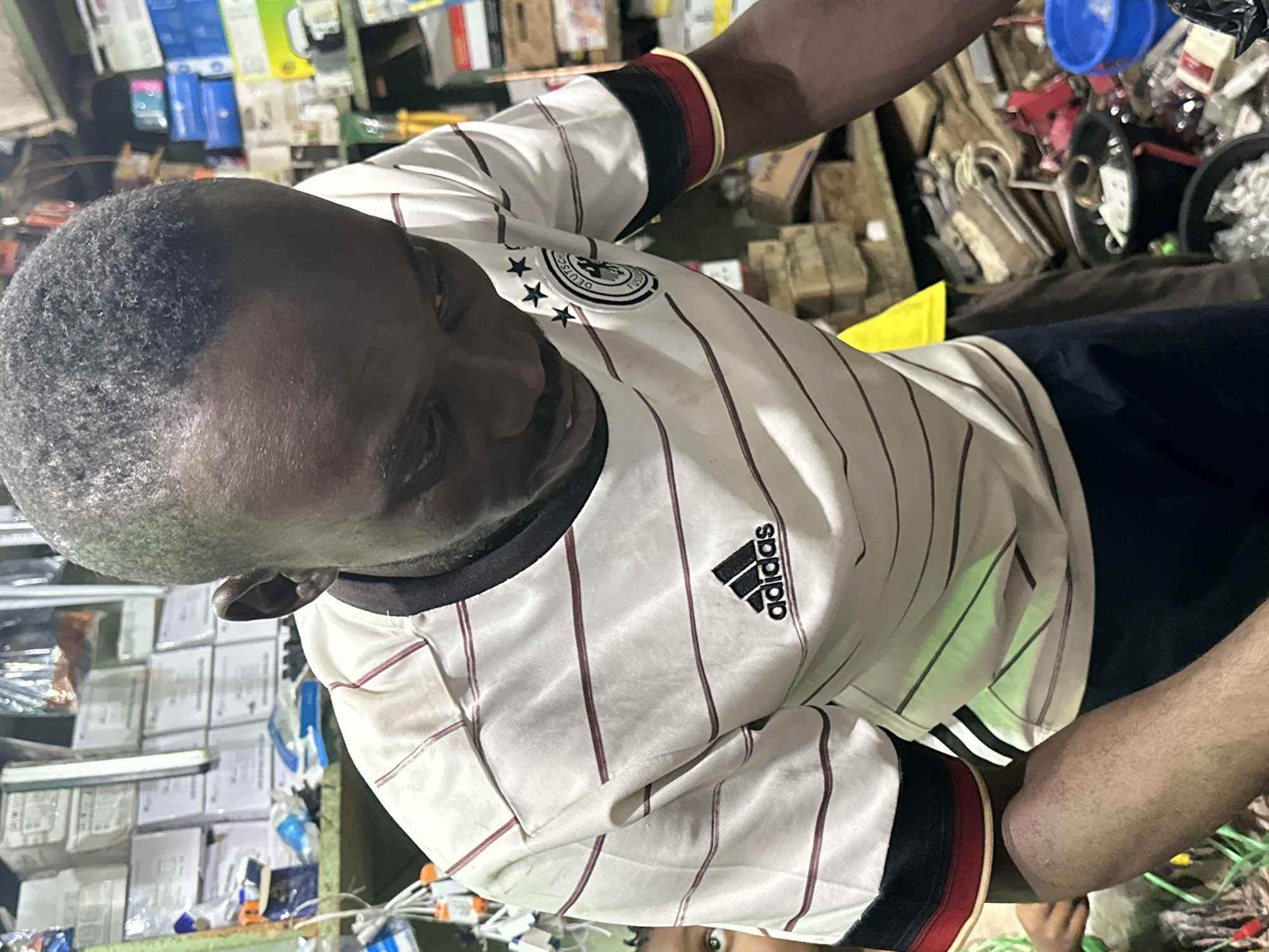 In Narayi, Nigeria, a man of African descent is captured in a medium shot, his head angled downwards as he appears to be engaged in an activity in a cluttered, indoor environment. He is wearing a white Adidas t-shirt with thin maroon horizontal stripes and black and red trim on the collar and sleeves. The shirt bears the Adidas logo and the emblem of the German national football team. His skin is dark, and his hair is short and textured. His facial expression is focused.
The background suggests a marketplace or workshop, filled with shelves stacked with various items, including boxes, electronic components, and what appear to be tools. Some shelves are also laden with merchandise in packaging. In the foreground, to the left of the man, a child's eye is visible, hinting at another presence in the scene. The overall atmosphere is one of a busy, possibly commercial setting. The lighting is somewhat dim, with a green hue visible on the man's shirt, likely from artificial lighting in the store.
