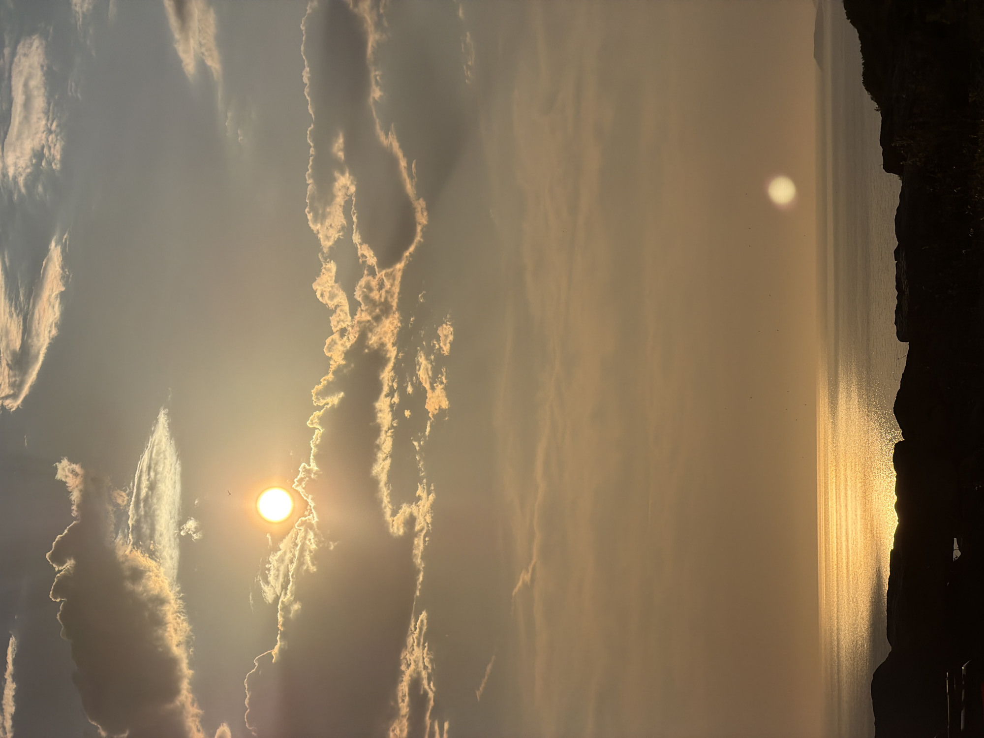 The image depicts a sunset over the Adriatic Sea, viewed from a high vantage point likely in or near Kovačko Polje, Montenegro. The sun, a bright orb, is partially obscured by clouds in the upper left quadrant of the frame, casting a golden glow across the sky and reflecting intensely on the water. The sea is calm, with the sunlight creating a shimmering pathway. In the distance, a hazy coastline is visible to the right. The foreground is dominated by dark, silhouetted hills or landmass, with hints of vegetation. A small, distant sun-like phenomenon is visible on the horizon over the water, possibly a reflection or another atmospheric effect. A few small birds are scattered across the sky. No people or specific structures are identifiable, contributing to a natural landscape scene.