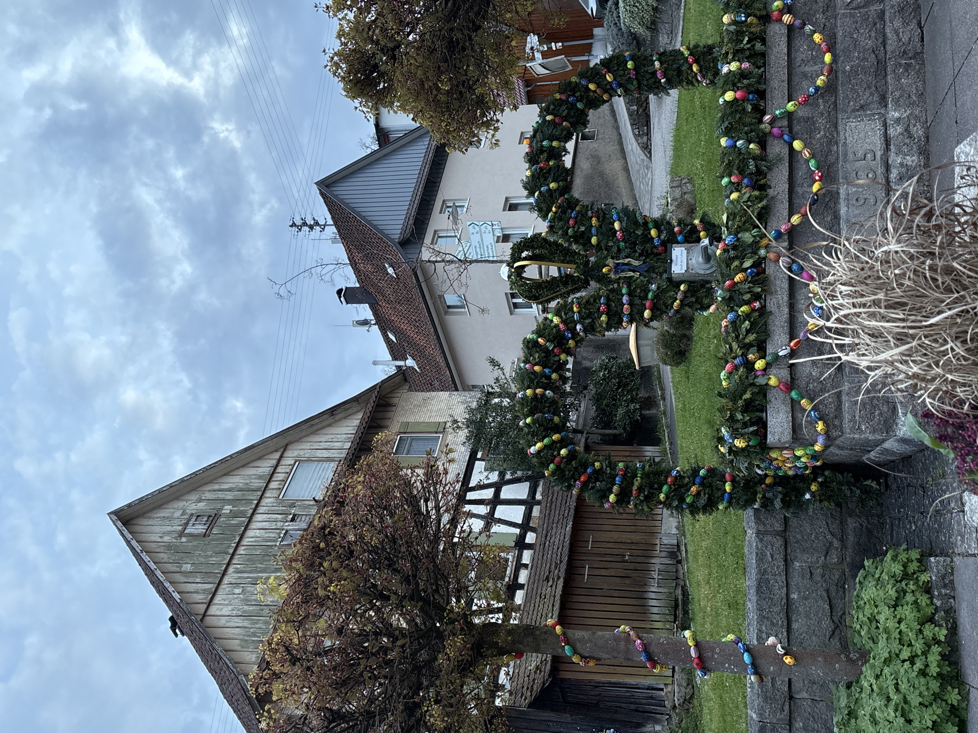 The image shows a street scene in Forbach, Germany, decorated for Easter. In the foreground, there is a stone wall with an Easter decoration made of greenery and colorful eggs. The decoration is draped over the wall and also around a wooden structure. Behind the wall, there is a green lawn, and beyond that, there are two buildings. The building on the left has a steeply pitched roof and exposed timber framing. The building on the right has a gray roof and a white facade. The sky is overcast and gray.