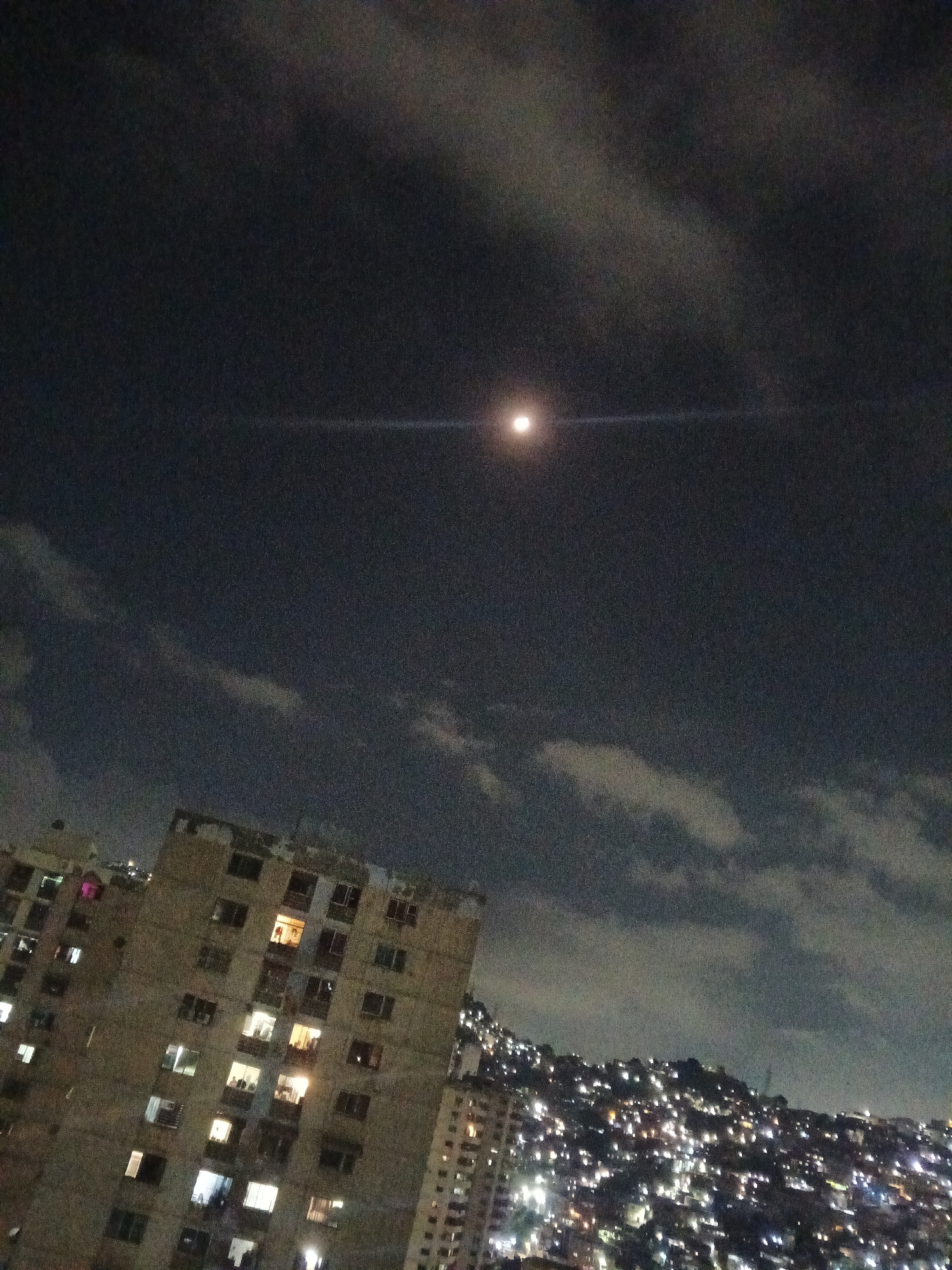 At night, a tall, beige building stands to the left of a densely populated hillside with numerous lights. A few windows in the building are lit. The sky is dark, with clouds and a bright moon with a lens flare extending horizontally. The city is Caracas, Venezuela.