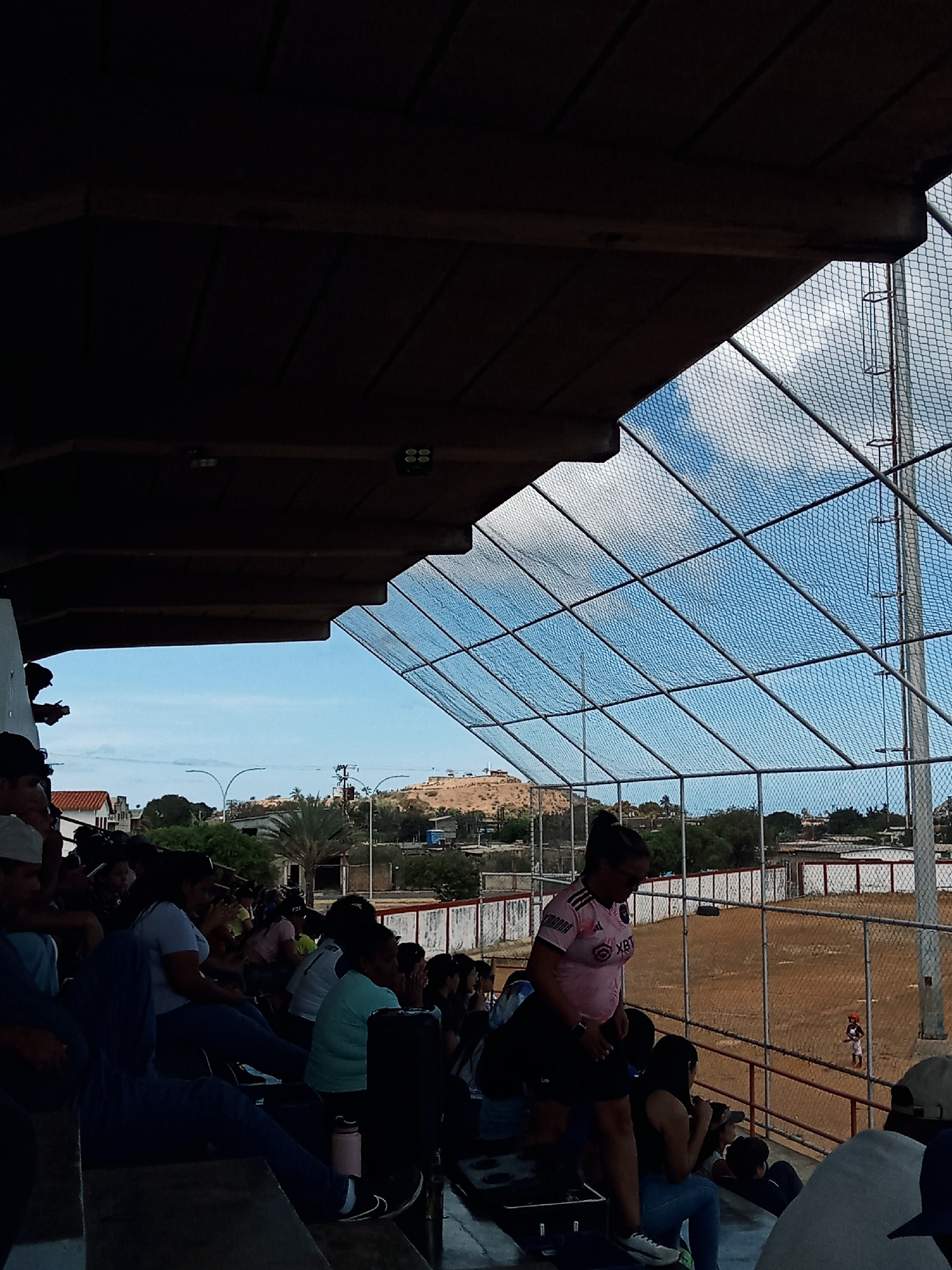 A crowd of people sits in the bleachers of a baseball stadium in Juan Griego, Venezuela. A chain-link fence separates the spectators from the dirt field where a baseball game is in progress. A woman in a pink shirt stands in the bleachers. Buildings, trees, and a small mountain are in the background. The sky is partially obscured by the roof of the bleachers and the netting.