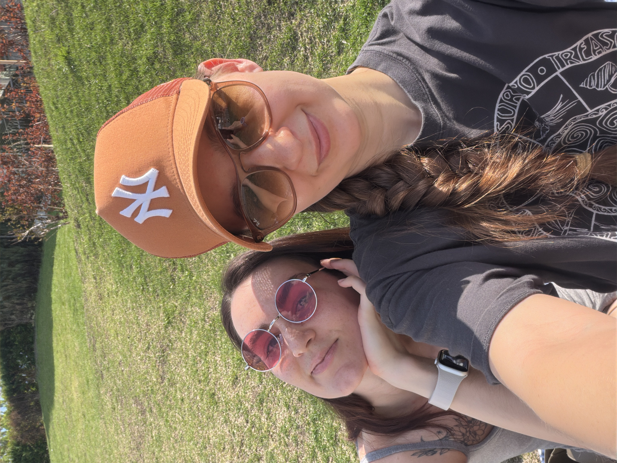 Two women are lying on a grassy lawn. The woman on top is wearing a brown baseball cap with a white "NY" logo, brown sunglasses, and a dark grey t-shirt with a white design on the front. Her hair is in a braid. The woman underneath is wearing round, pink-tinted sunglasses and has her hand on her face. She is wearing a grey tank top and a light-colored watch on her wrist. The background shows green grass and some trees and bushes. The location is outdoors on a sunny day, but the specific location in Tyumen, Russia cannot be determined from this image.