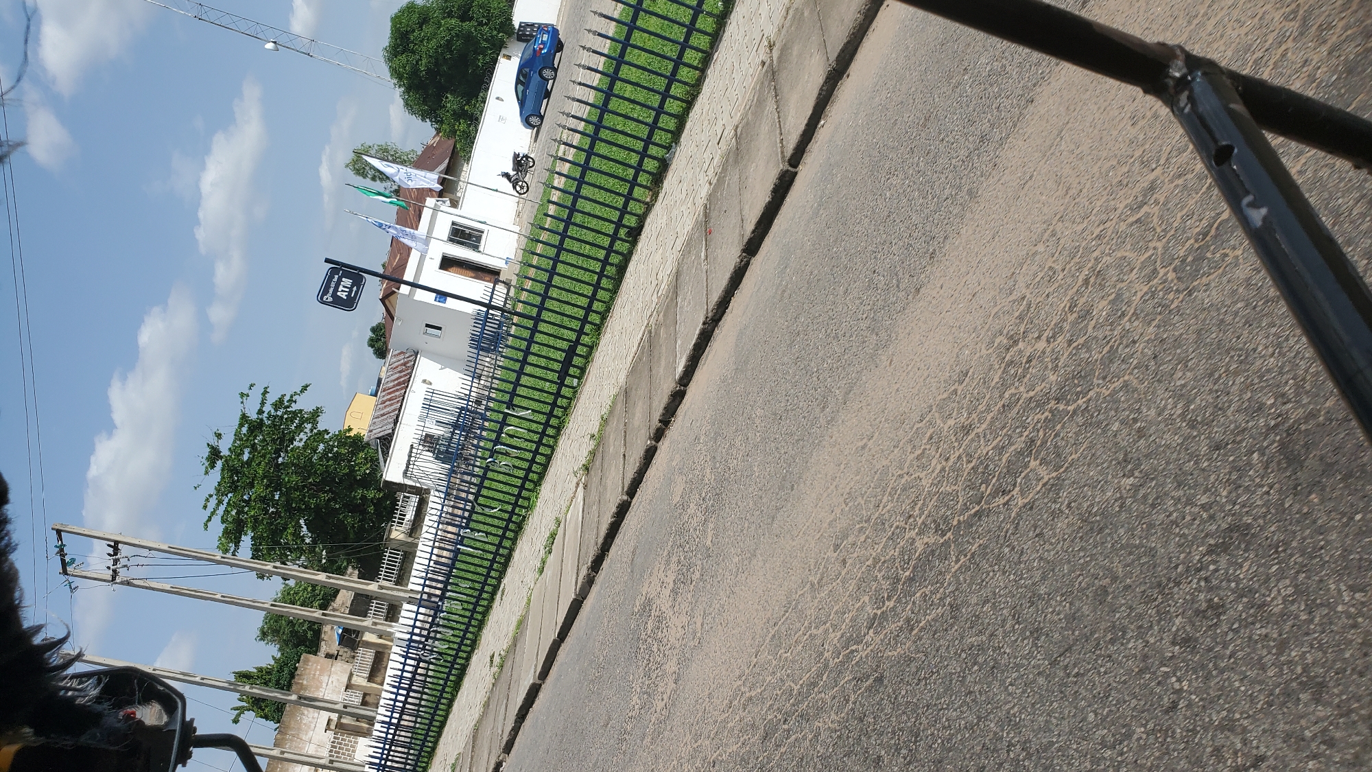 The image, taken in Jalingo, Nigeria, shows a street scene with a building featuring an ATM. A blue sedan is parked in front of the building, and a motorcycle is parked near the entrance. The building is enclosed by a dark metal fence, beyond which is a well-maintained green lawn. Flags are displayed outside the building. A power pole with electrical lines is visible in the background. The foreground is dominated by a paved road with a concrete sidewalk. The sky is clear with scattered white clouds. The perspective suggests the photo was taken from a moving vehicle.