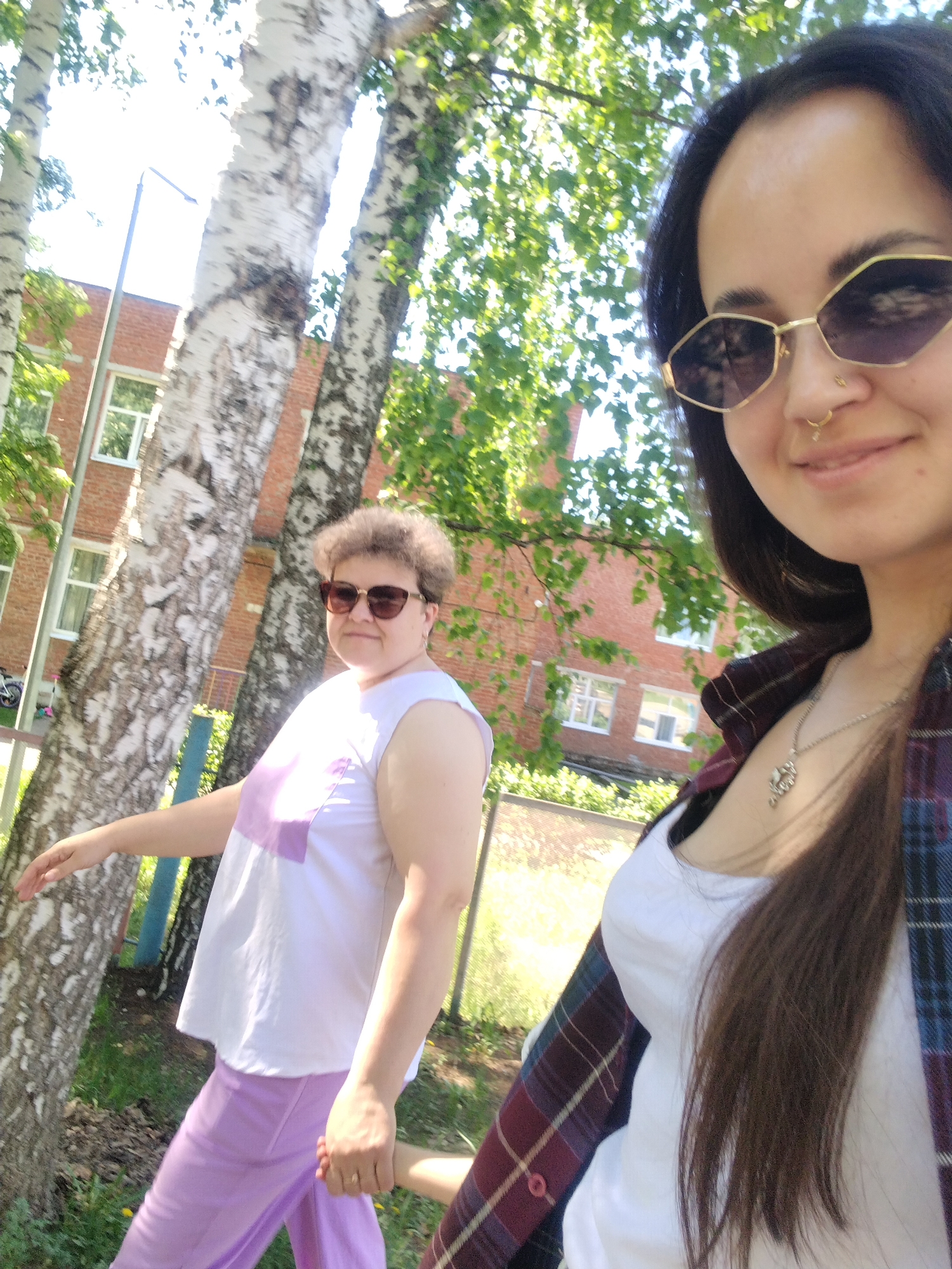 The image depicts two women walking together outdoors in Noviy, Russia. The woman on the right is closer to the camera, wearing sunglasses, a white top, and a partially visible plaid shirt. She has long, dark hair and a nose ring. The woman on the left, further in the background, is wearing sunglasses, a white top with a purple square design, and purple pants. They appear to be holding hands. The scene is outdoors, with birch trees and a red brick building visible in the background. A streetlight and a partial view of a parked vehicle are also visible. The scene is well-lit, suggesting it is daytime.