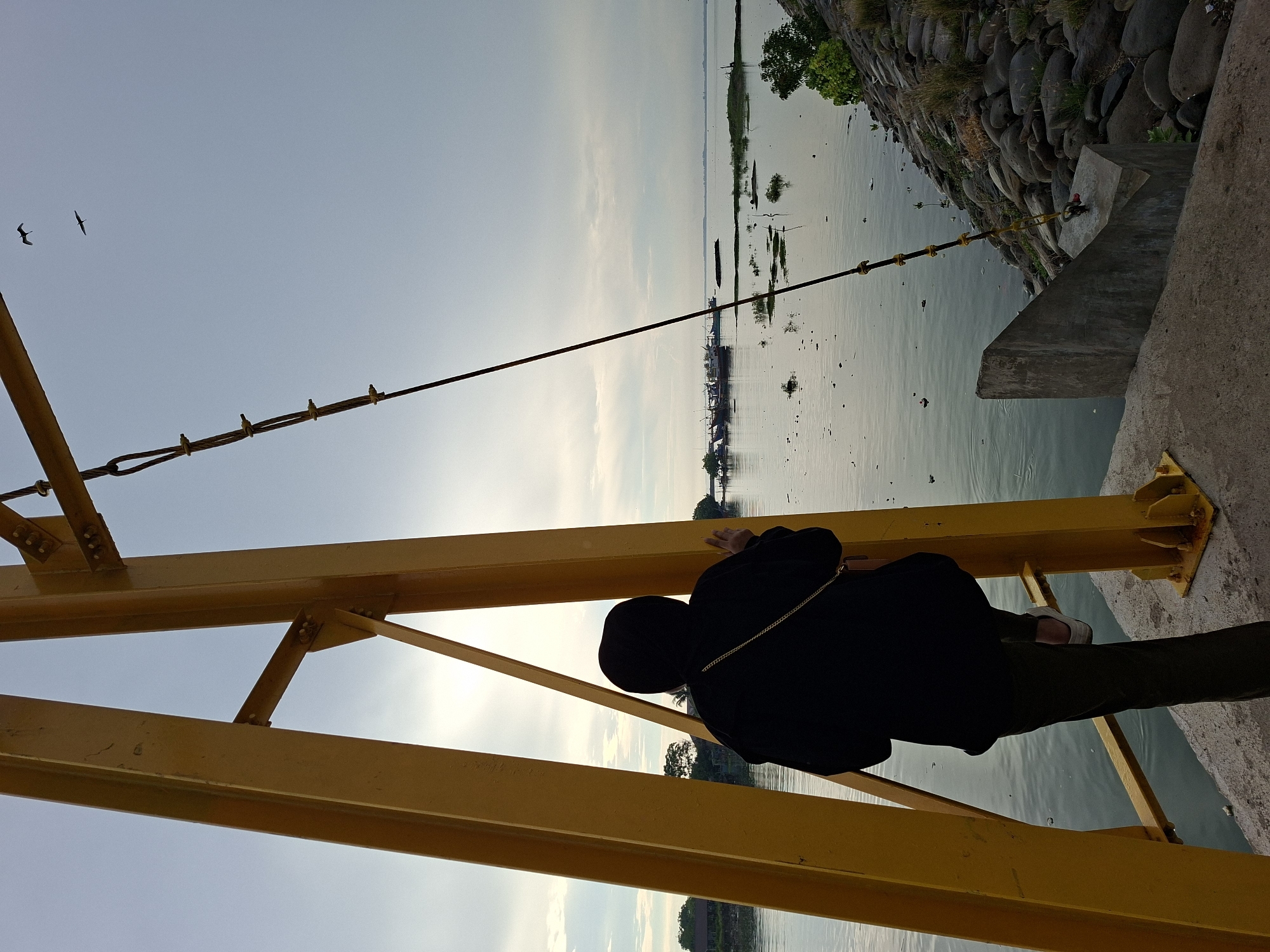 In Lhokseumawe, Indonesia, a person clad in a black hijab and jacket stands with their back to the viewer, one foot on a rung of a bright yellow metal structure that appears to be part of a crane or industrial equipment. The person is positioned to overlook a body of calm water where several boats are docked in the distance. The sky is overcast with a hint of sunlight breaking through. The foreground includes a concrete pier with a rocky embankment to the right. Two birds are visible in flight against the sky. The overall scene suggests a waterfront industrial or port area.