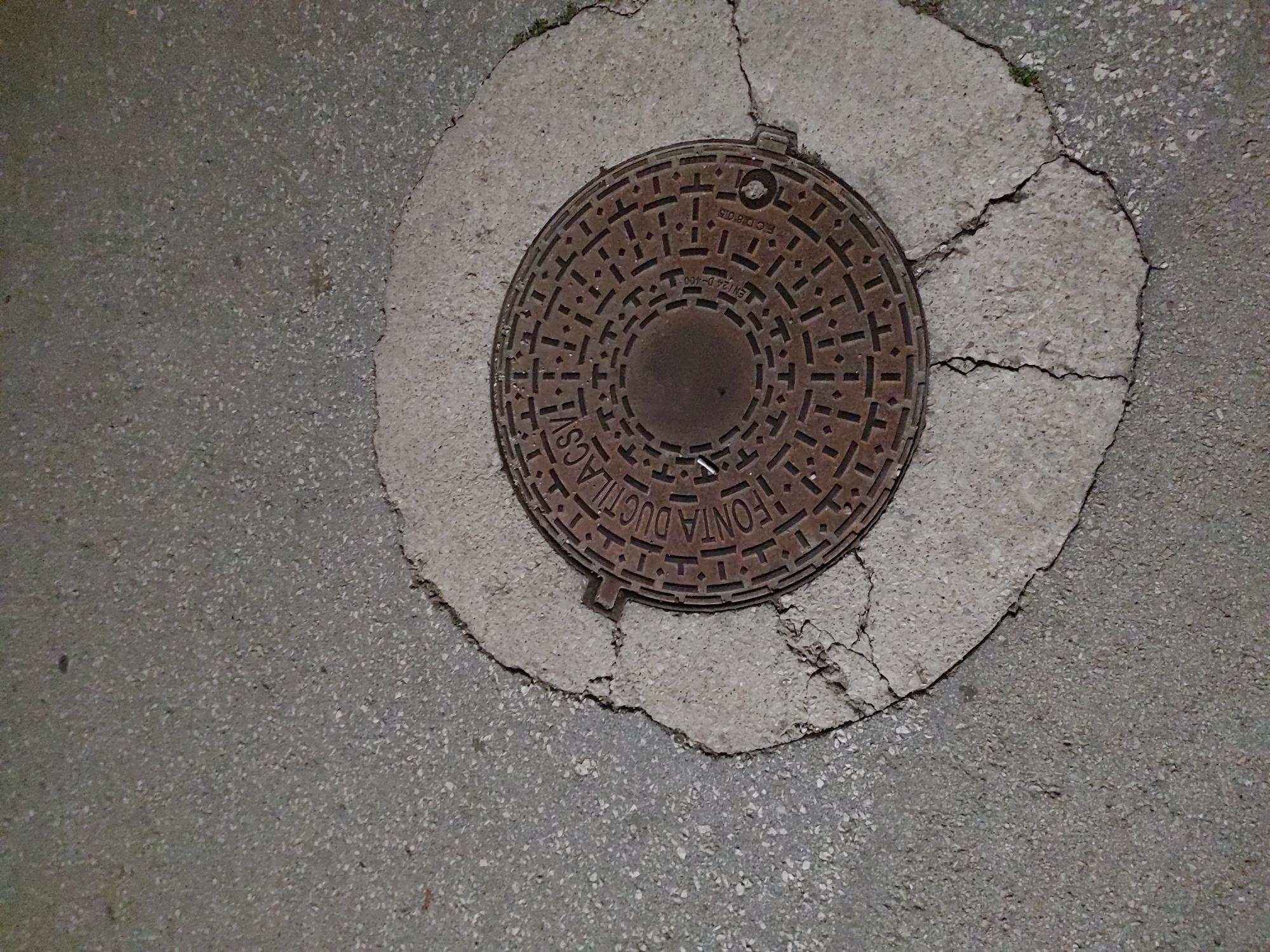 The image shows a manhole cover embedded in a cracked concrete surface, which is surrounded by asphalt. The manhole cover is circular and made of a dark, textured metal, with intricate patterns and some inscribed text that appears to include "ROMA DUCTILACSV" and "EN124 D-400". The concrete around the manhole cover is light gray and shows significant cracks radiating outwards, suggesting wear and tear. The asphalt is a darker gray and has a granular texture. There are no people or animals visible in the image, nor any specific identifiable landmarks. The lighting suggests it might be nighttime or an overcast day, casting subtle shadows. The scene is an urban street or sidewalk environment. This image does not contain enough information to determine its location in Tervel, Bulgaria.