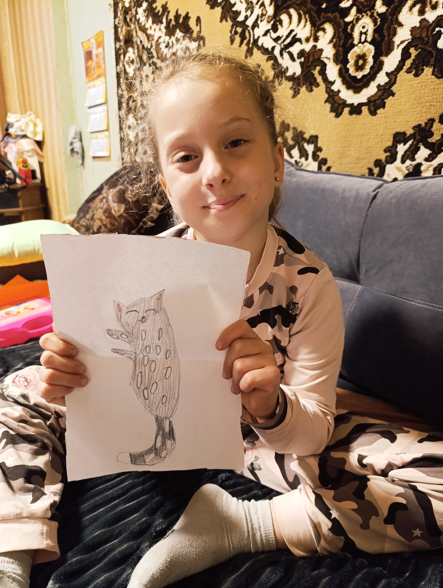 A young girl is sitting on a couch, holding up a drawing of an animal. The drawing depicts a grey animal with black spots, a smiling face, and small arms. The girl has blonde hair and is wearing a light pink pajama set with a camouflage pattern. She is smiling slightly. She is sitting on a dark blue couch with a textured blanket. Behind her, there is a brown and beige patterned tapestry on the wall. There is also a wooden cabinet with items on it and a stack of calendars on the wall. The location is in an indoor space, likely a home in the city of Unknown, Unknown.