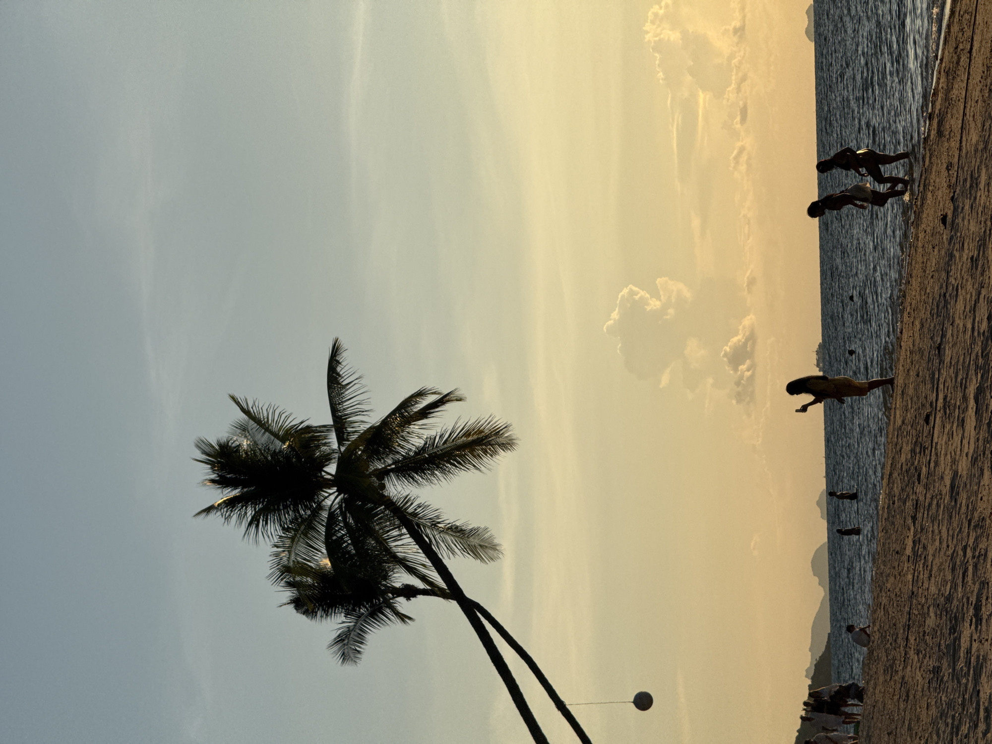 This image depicts a beach scene in Baan Tai, Thailand, during sunset. Two palm trees are silhouetted against a gradient sky of pale blue to warm yellow. A round, dark object hangs from a rope attached to one of the palm trees. In the foreground, a sandy beach stretches towards the sea. Several people are visible on the beach and in the shallow water. On the right, two figures walk near the water's edge, and further to the left, a lone figure stands holding a phone, possibly taking a photo. More individuals are scattered along the shoreline and in the distance, some wading in the ocean. The sea is dark and reflects the sky's colors. In the background, islands or landmasses are visible on the horizon, with a ship also present in the distance. The overall atmosphere suggests a peaceful evening on a tropical beach.