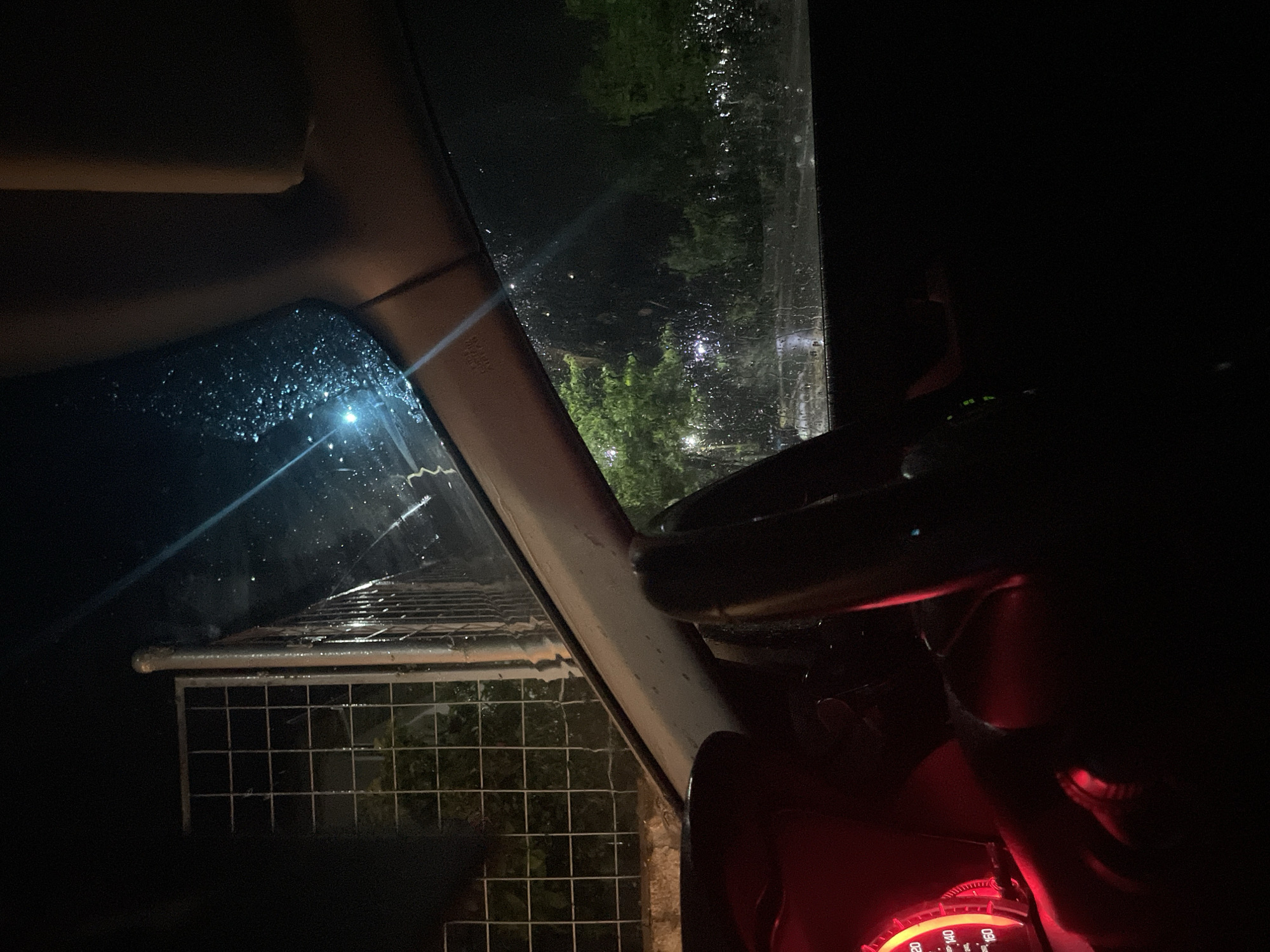 The image shows the view from inside a car on a rainy night. The dashboard and steering wheel are visible, illuminated by a red glow from the instrument panel. Through the rain-streaked windshield, a fence and some trees are visible. The location is unknown. No people are visible.