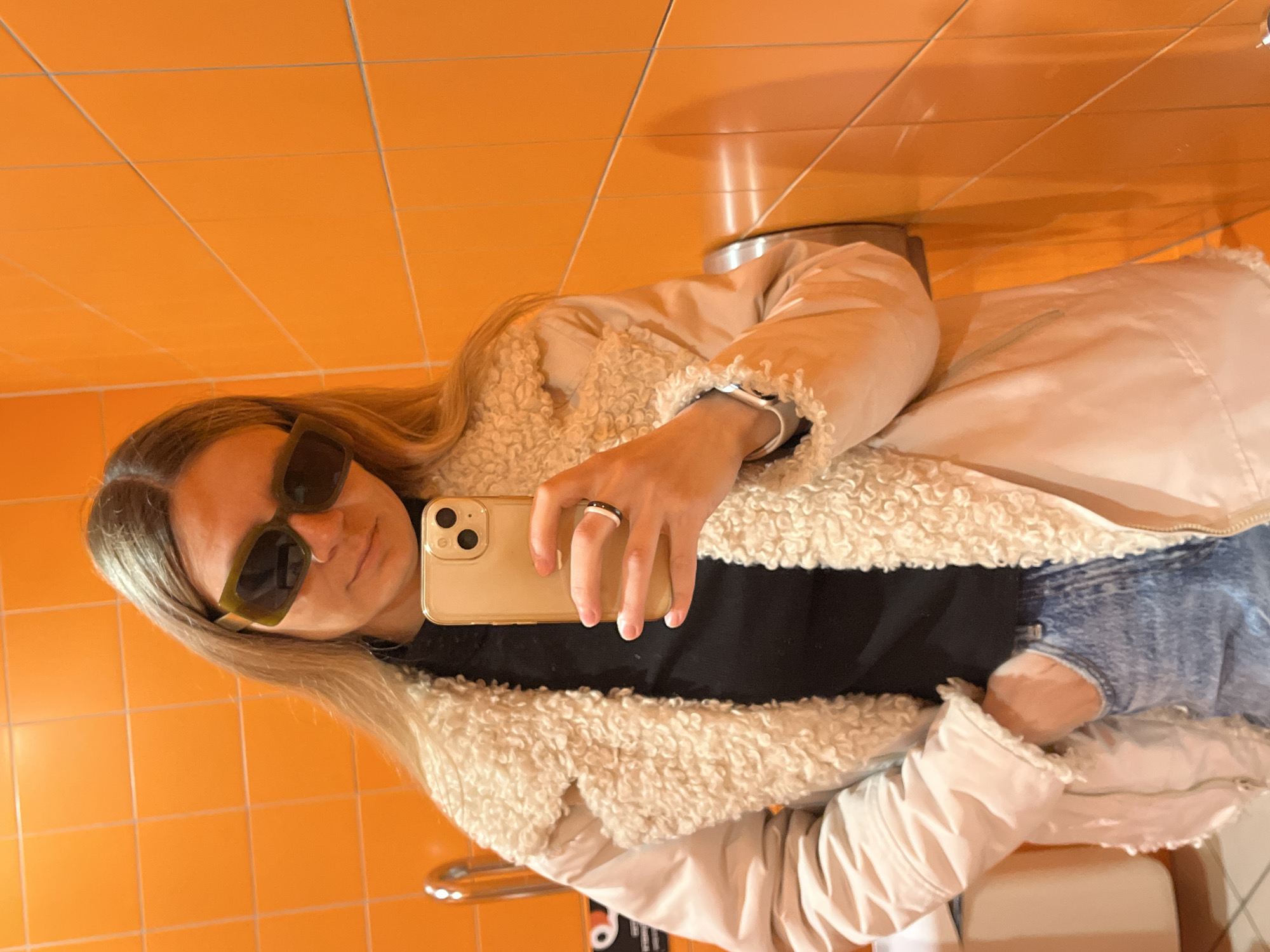 A young woman is taking a selfie in a bathroom with orange tile walls. She is wearing sunglasses, a black top, jeans, and a light-colored jacket with a furry collar. She is holding a gold iPhone in her right hand. The location may be the city of Chaikovsky, Russia.