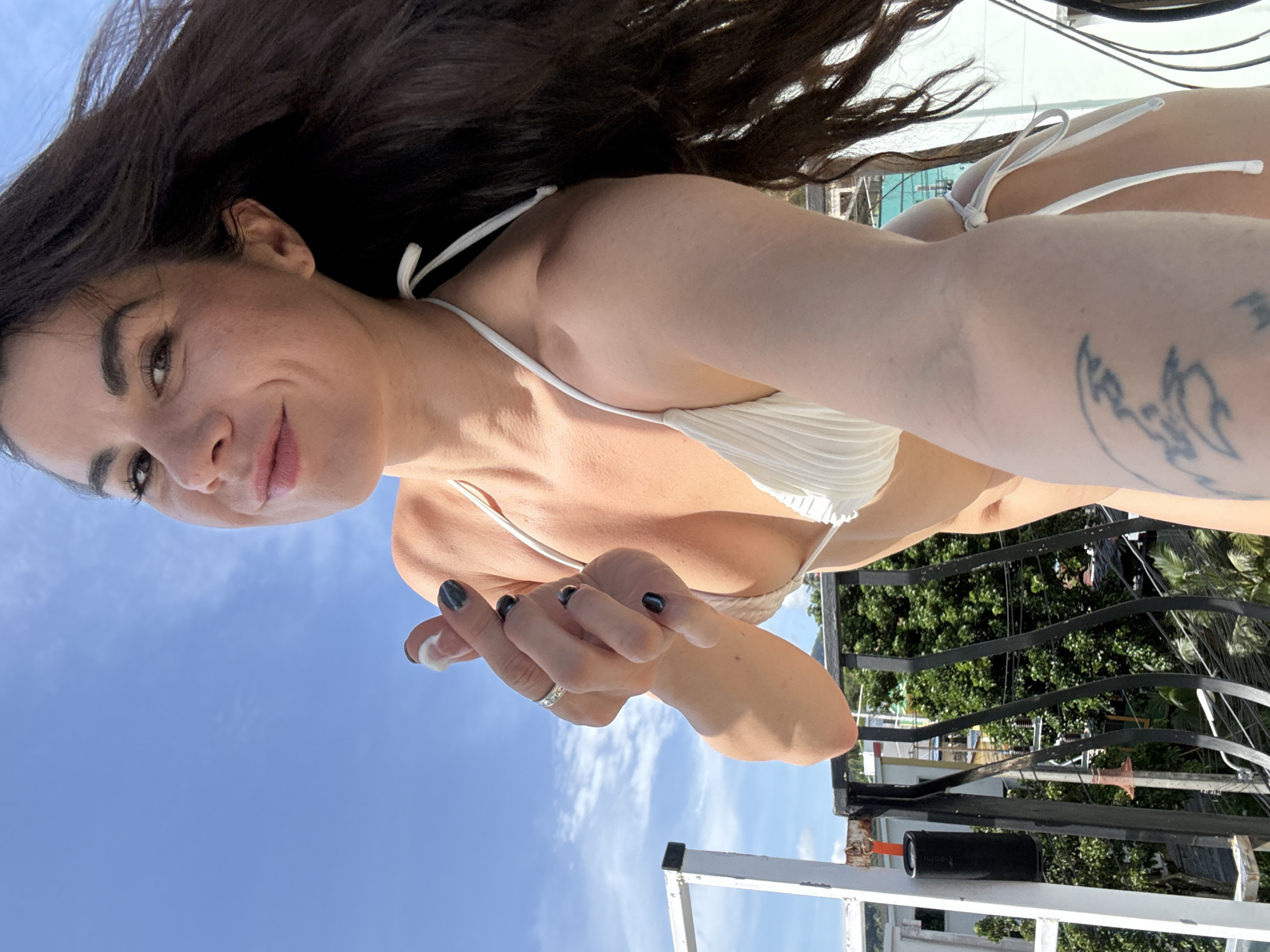 A woman in a white bikini poses for a selfie on a balcony overlooking a city with lush green trees and buildings under a clear blue sky. Her right hand is raised, forming a finger heart gesture, and she wears a silver ring on her left ring finger. Her long, dark hair is windswept, framing her face as she smiles. A small black speaker with an orange loop is attached to the white railing of the balcony.