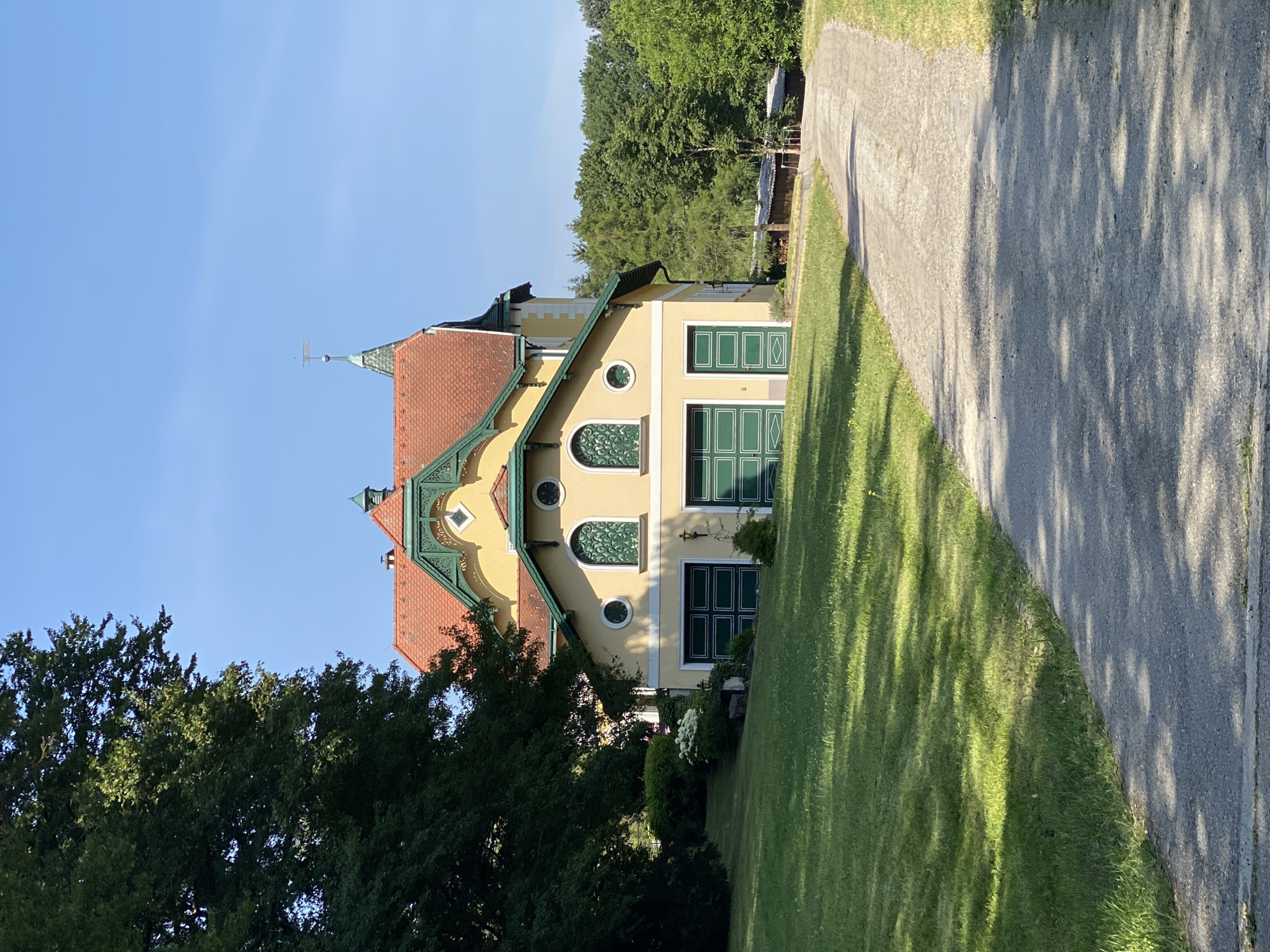 The image captures a sunny day in Pressbaum, Austria, focusing on a light yellow, multi-story building with a red tiled roof and green decorative accents. The building has a distinctive architectural style with several arched windows featuring intricate green lattice work. Two large green garage doors are visible on the lower level, along with a smaller green door. The building is set back from a gravel driveway that curves through a well-maintained green lawn. To the left of the house, a large, mature tree with dense foliage casts shadows on the lawn. In the background, a dense line of trees creates a natural backdrop, with a clear blue sky above. The scene appears to be a residential or possibly a historic property, with no people or animals visible.