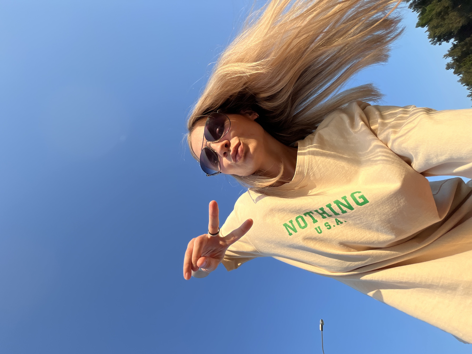 A woman in sunglasses and a beige "NOTHING U.S.A." sweater is making a peace sign in front of a clear blue sky, possibly on a sunny day in Chaikovsky, Russia. The location is inferred from the presence of a possible street light pole in the lower center of the image and foliage from trees in the upper right. The woman's blonde hair is blowing upwards.
