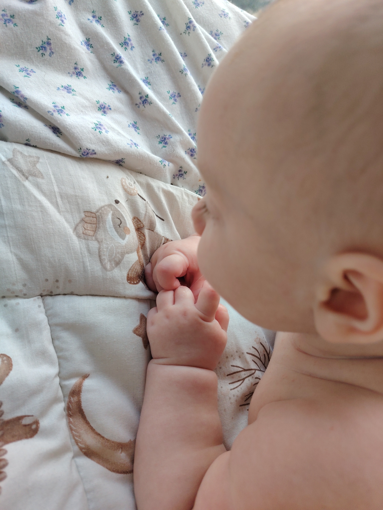 The image, taken in Konakovo, Russia, is a close-up shot of a baby lying on a soft, quilted surface. The surface has a pattern of small purple flowers on a white background in the upper portion of the image, and a pattern of brown animal figures and a crescent moon on a cream-colored background in the lower portion. The baby's head, shoulder, and arms are visible, and they are gripping their own hand. The baby's skin is fair and smooth. The lighting is soft and natural, suggesting an indoor setting. The overall impression is one of tenderness and innocence.