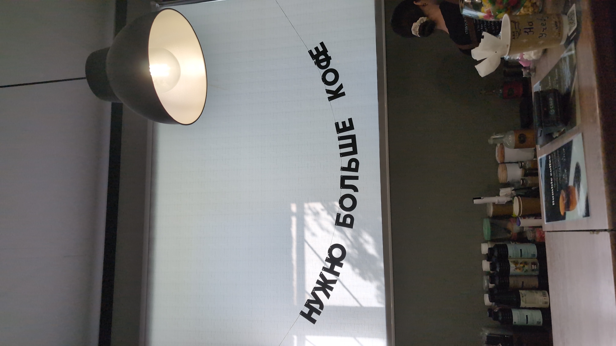 The image captures a cafe interior in Bor, Russia. The primary subject is a wide shot of a counter area and a wall adorned with a banner. On the left side of the frame, a large, dome-shaped pendant lamp with a bright bulb hangs, casting a warm light. To its right, a white roller blind covers a window, with shadows of leaves or blinds visible on it.
A banner with black, block letters is strung across the blind, reading "НУЖНО БОЛЬШЕ КОФЕ" which translates to "MORE COFFEE NEEDED." The letters are arranged in a curve, with "НУЖНО" on the left, arching upwards towards "КОФЕ" on the right.
On the right side of the image, the counter displays various items. A person, mostly out of focus and facing away from the camera, is visible near the top right. In front of them are several objects, including what appear to be small cups, bottles, and possibly jars of candy or condiments. One cup, in the upper right, has handwritten text that is partially legible. Below this, a collection of bottles and cups are neatly arranged on shelves. A small sign with the text "Bumble coffee" is visible on the counter.
The overall impression is a casual, possibly bustling, cafe environment. The lighting suggests it could be daytime, with the shadows on the blind indicating ambient light from outside. The setting appears to be indoors, likely a modern cafe.