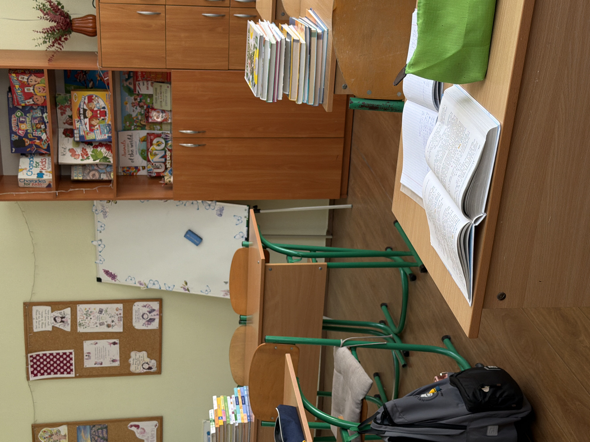 The image shows a classroom setting with a focus on several student desks. A stack of books sits on the edge of one desk, while an open book and a green bag rest on another. A grey backpack is placed on the floor beside the desk. In the background, wooden cabinets, bookshelves filled with games and books, and a whiteboard adorned with butterfly decorations are visible. A bulletin board with displayed items is affixed to the wall. The room features green desk and chair frames. The location appears to be a classroom.