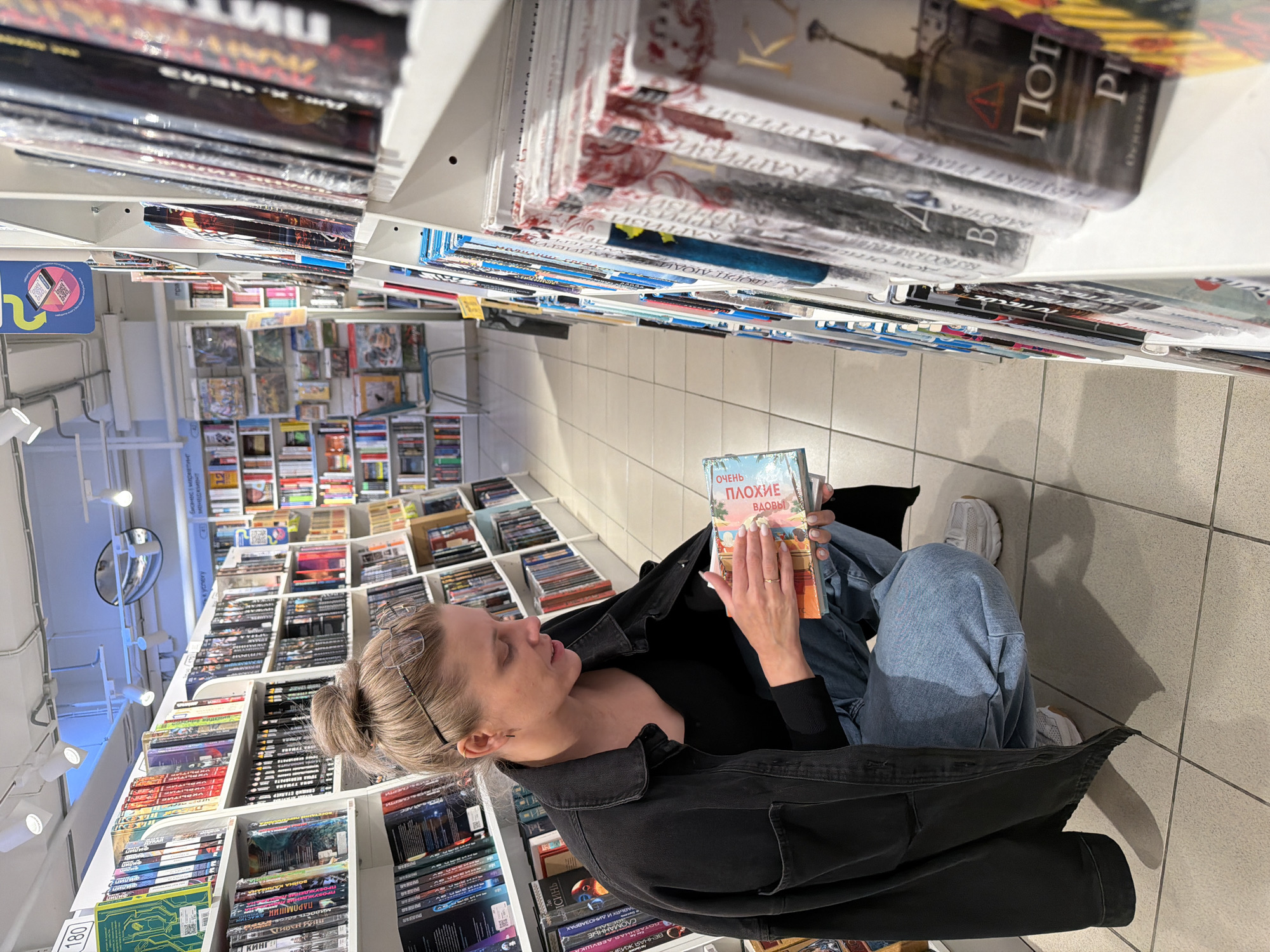 A woman is sitting on the floor in a bookstore aisle, reading a book with the title "Very Bad Widows" written in Russian. She is wearing a black jacket, blue jeans, and white sneakers. Her hair is pulled back in a bun, and she has glasses on her head. The aisle is lined with shelves of books, and there are other customers browsing in the background. Some of the books on the shelves have titles that appear to be written in Russian. Based on the store signage, it is in Izhevsk, Russia.